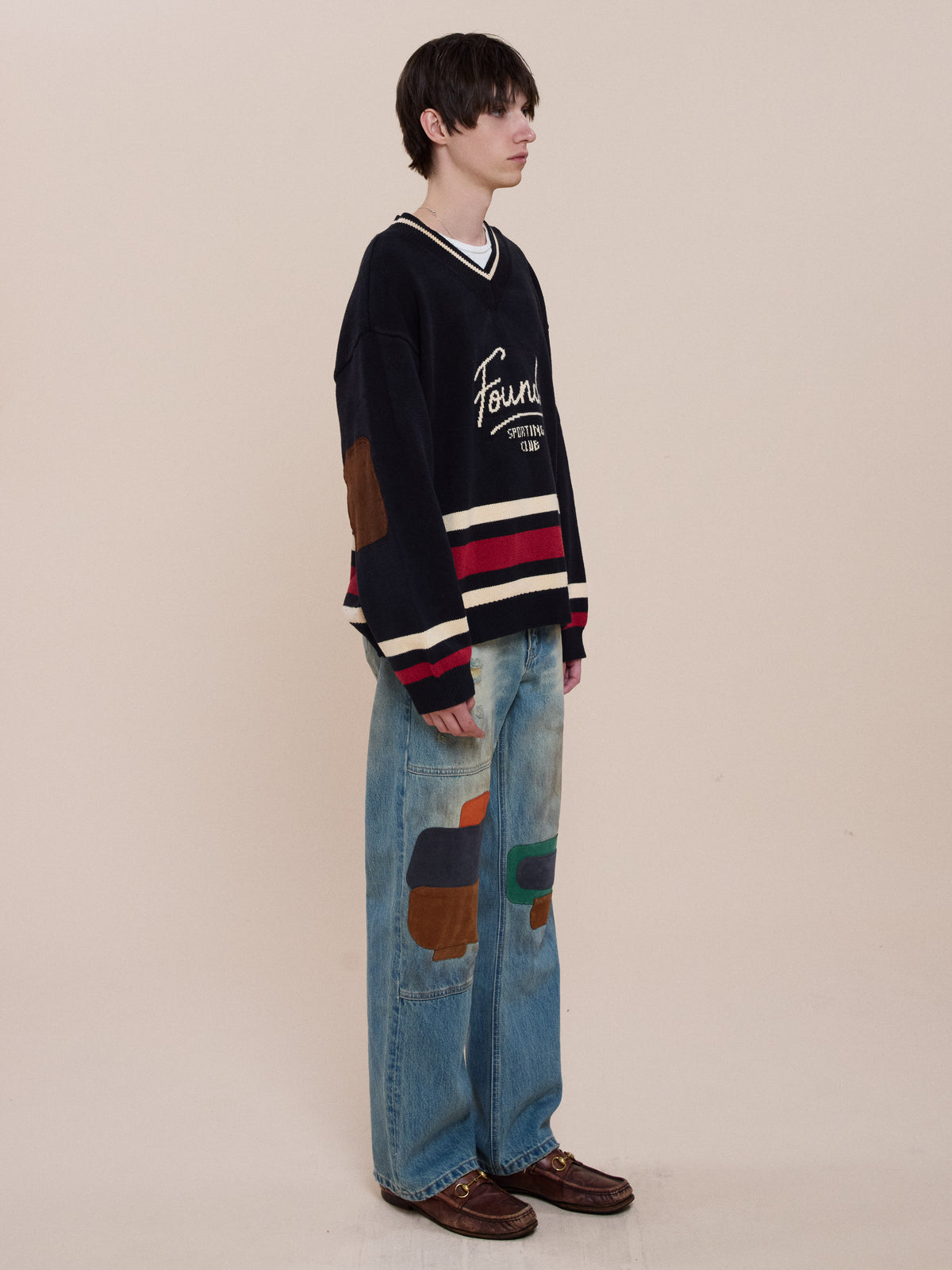 A person stands sideways in a FOUND Sport Club Varsity Sweater, baggy jeans with colored patches, and brown shoes against a plain beige background.