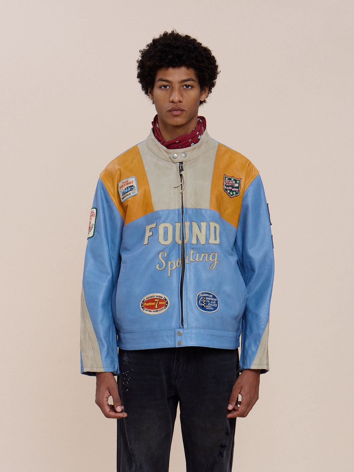 A person faces forward in a Sport Racer Jacket by FOUND, featuring racing patches and “FOUND Sporting” text, paired with black pants against a plain background.