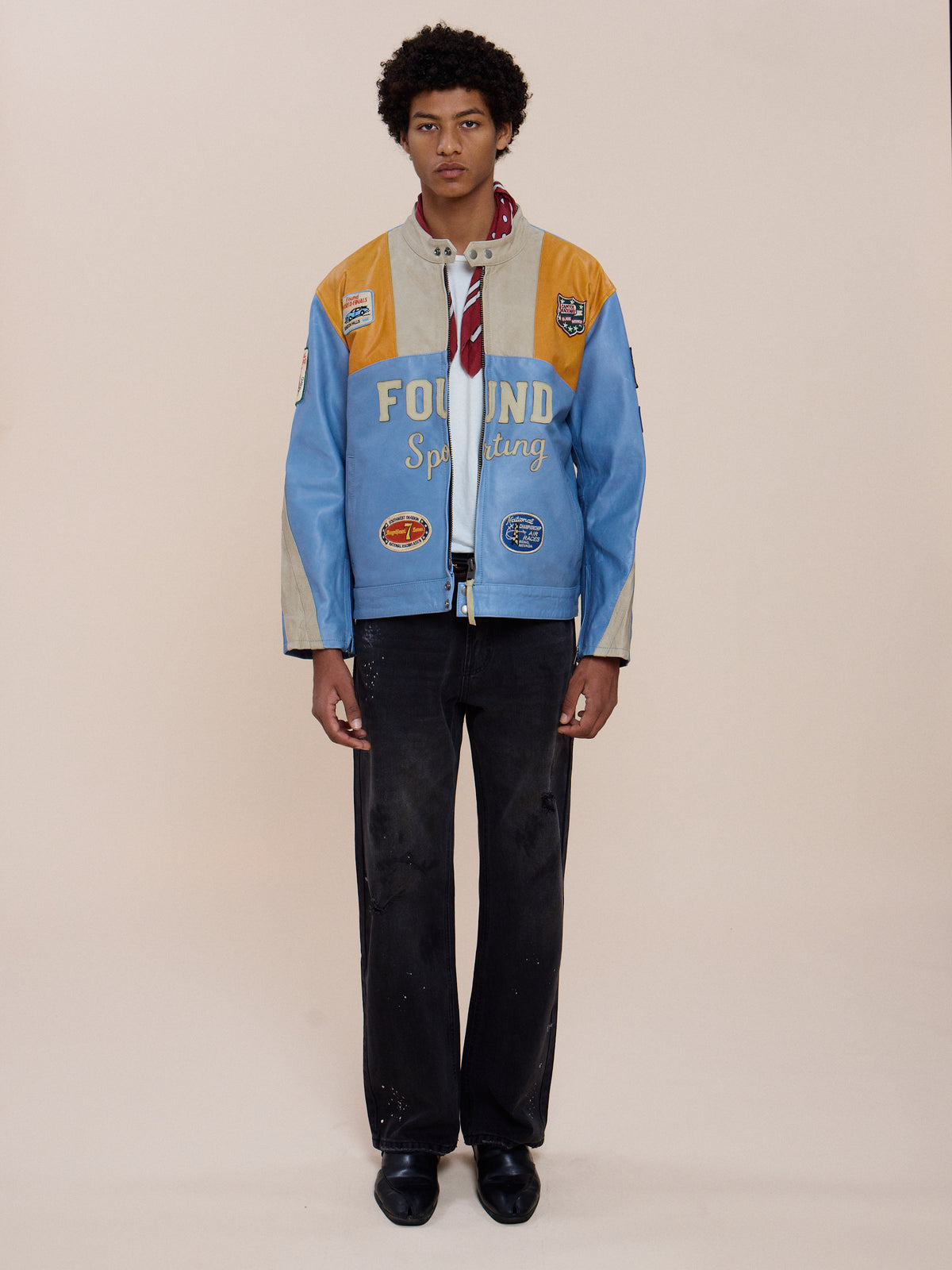 A person wears the FOUND Sport Racer Jacket in light blue and yellow over a shirt with a red scarf, black pants, and black shoes against a plain beige background.