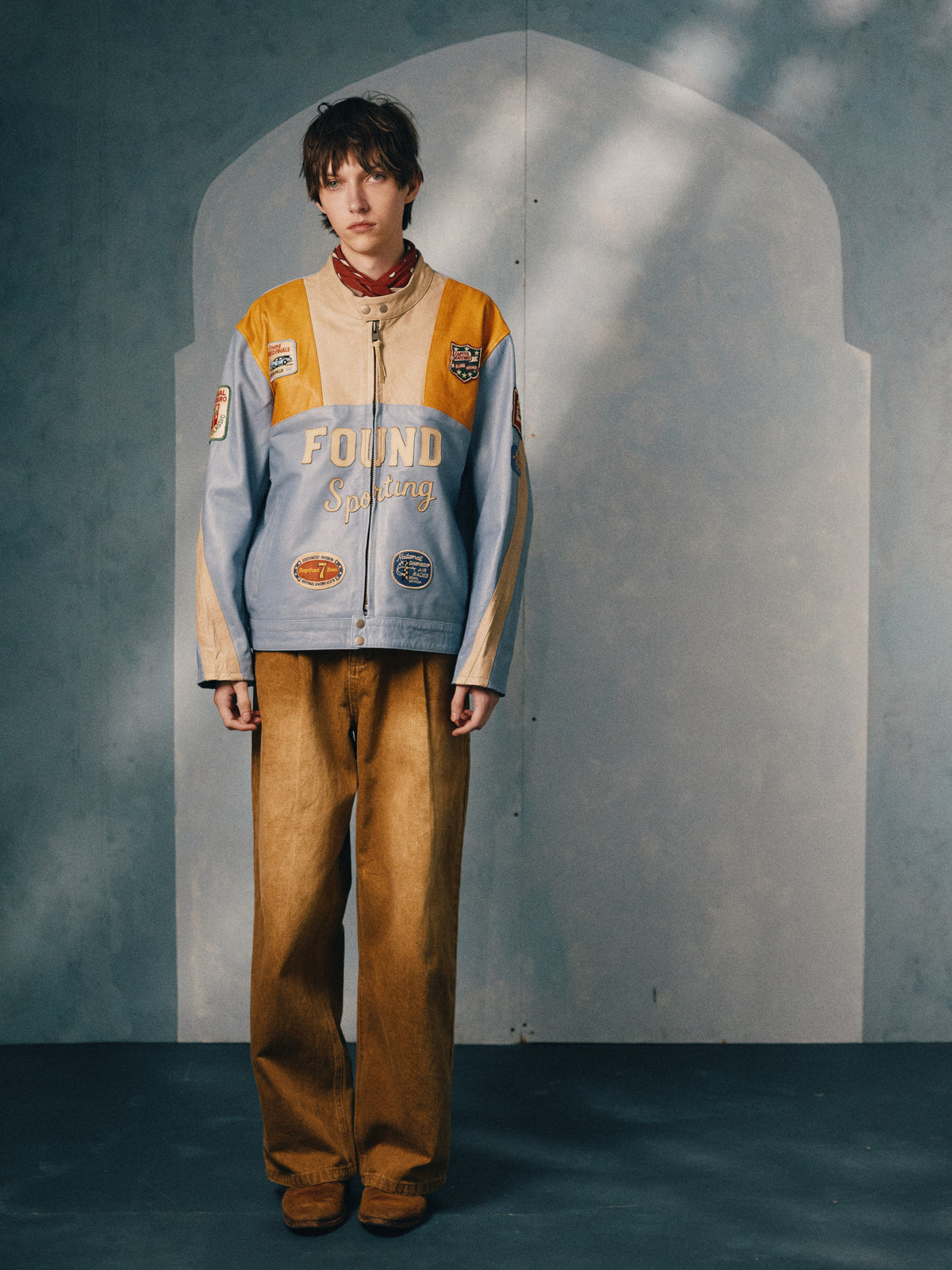 Person indoors in a blue and yellow Sport Racer Jacket by FOUND, paired with brown wide-leg pants and a red scarf, standing against a gray wall with an arched shadow.