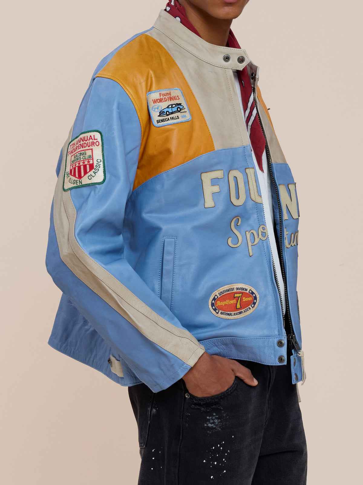 Person wearing the FOUND Sport Racer Jacket—blue, orange, and beige with racing-inspired patches—over a red and white shirt and black pants.