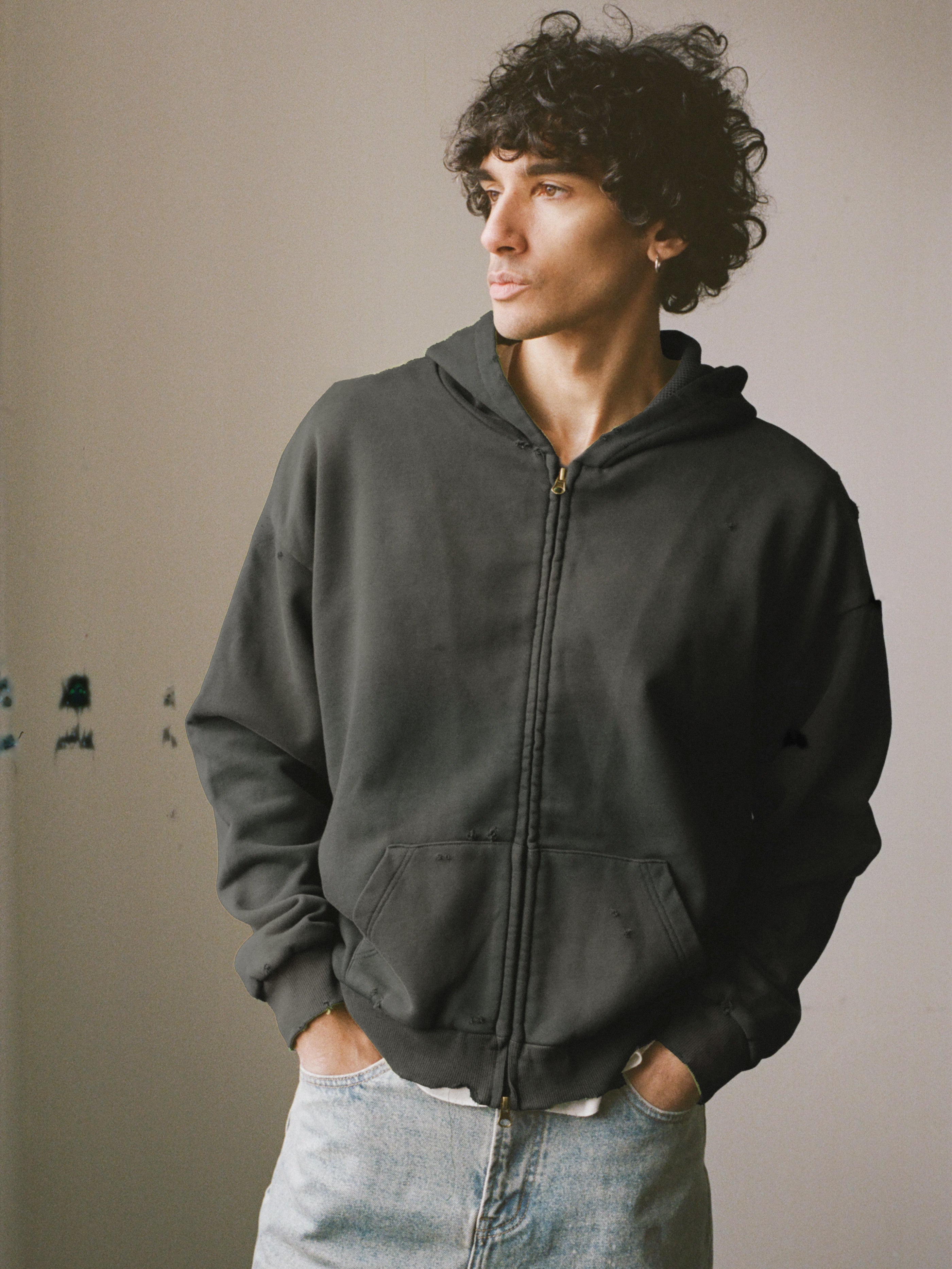 A person with curly hair, hands in pockets, stands indoors wearing the FOUND Distressed Thermal Lined Zip Up Hoodie and light blue jeans, looking to the side.