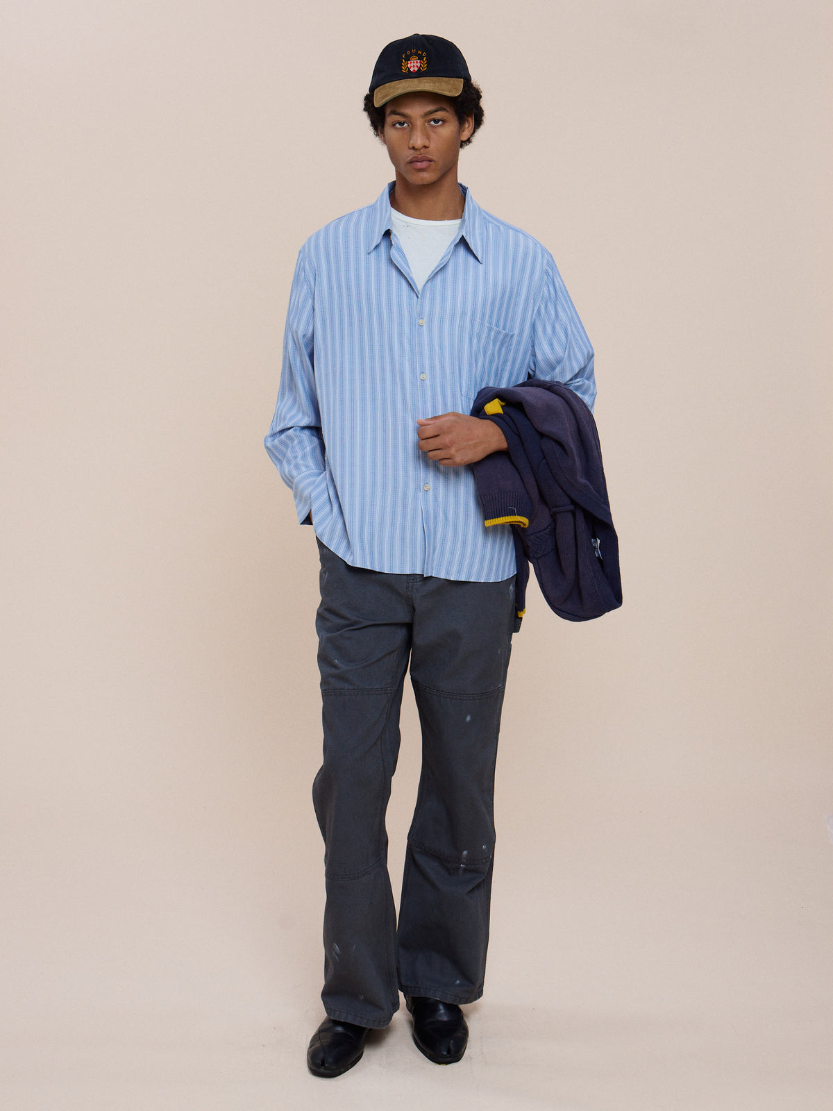 A person wears a FOUND Stripe Pocket Oxford Shirt, gray pants, black shoes, and a black cap, holding a jacket over their left arm against a plain background.