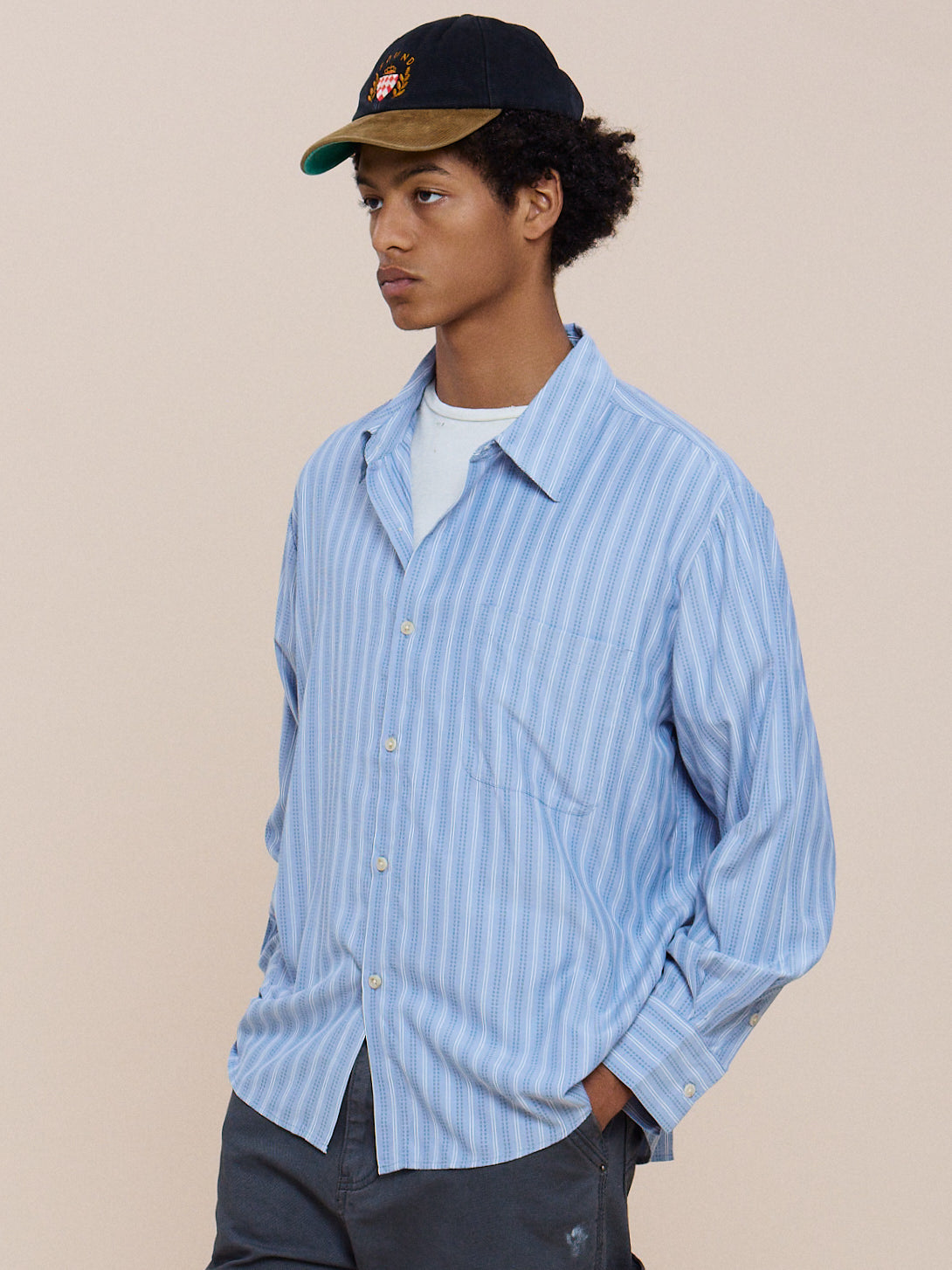 A person models the FOUND Stripe Pocket Oxford Shirt in a unisex relaxed fit, layered over a white tee with gray pants and a black cap against a beige background.