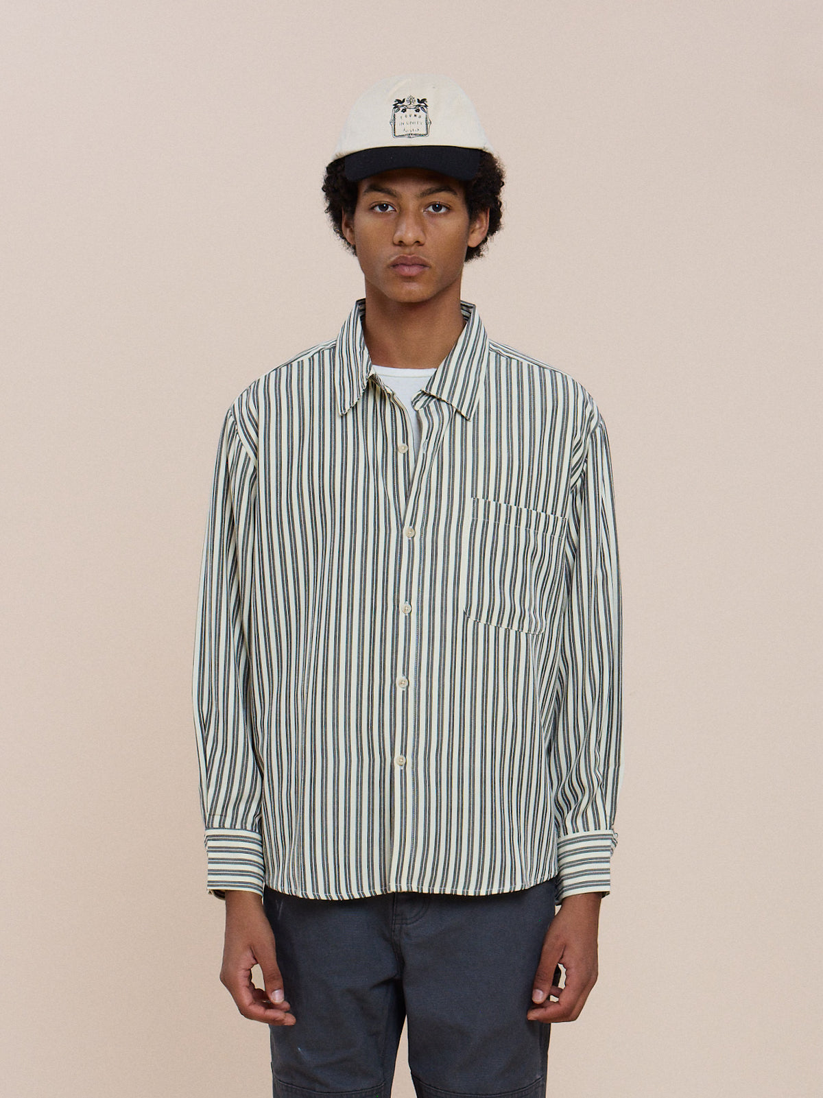 A person in a FOUND Stripe Pocket Oxford Shirt, white baseball cap, and dark pants stands against a plain light background.