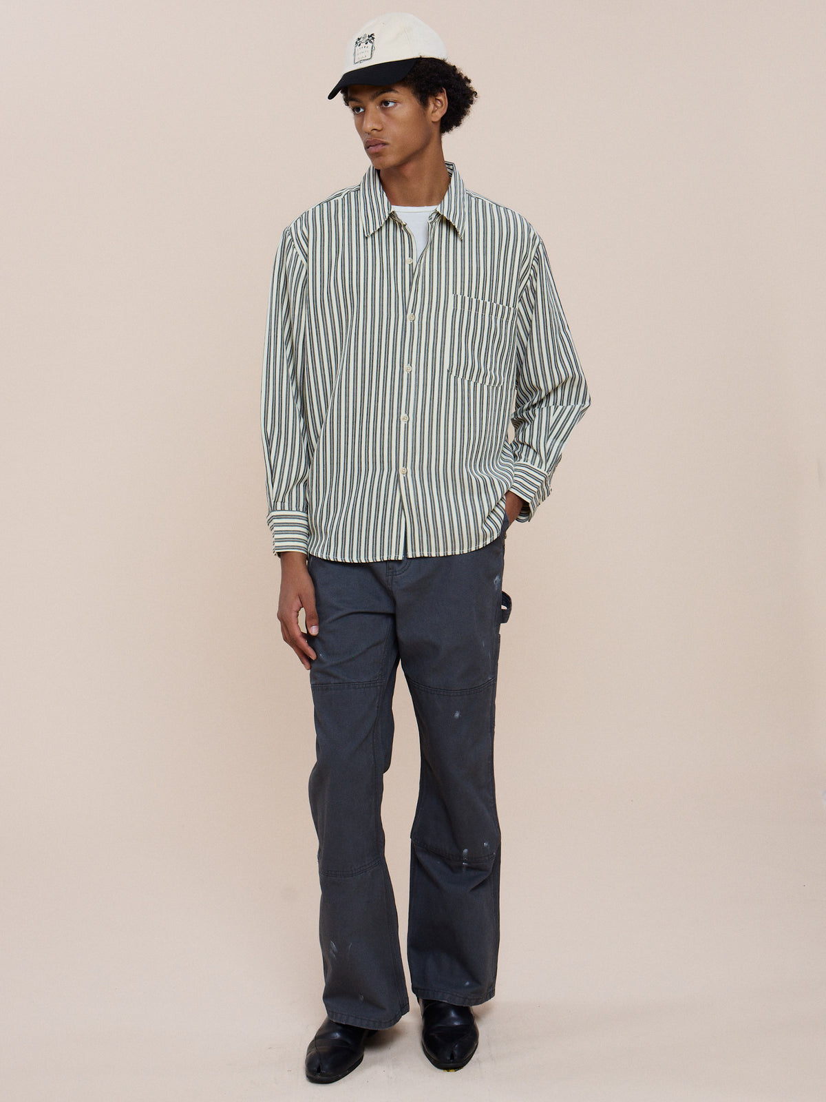 Person stands against a plain background wearing a white cap, gray paint-mark pants, black shoes, and FOUND Stripe Pocket Oxford Shirt in green and white vertical stripes.