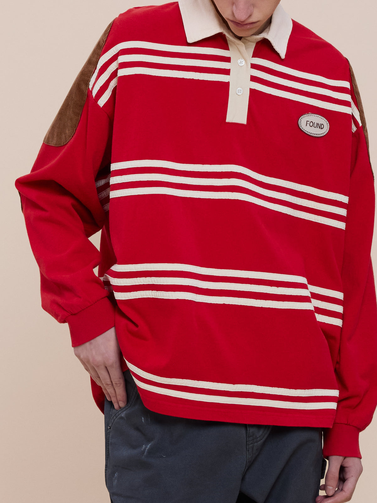 Person wearing the FOUND Striped Corduroy Rugby Polo, featuring red and white stripes, tan collar, brown elbow patches, and looking down with one hand in a pocket.