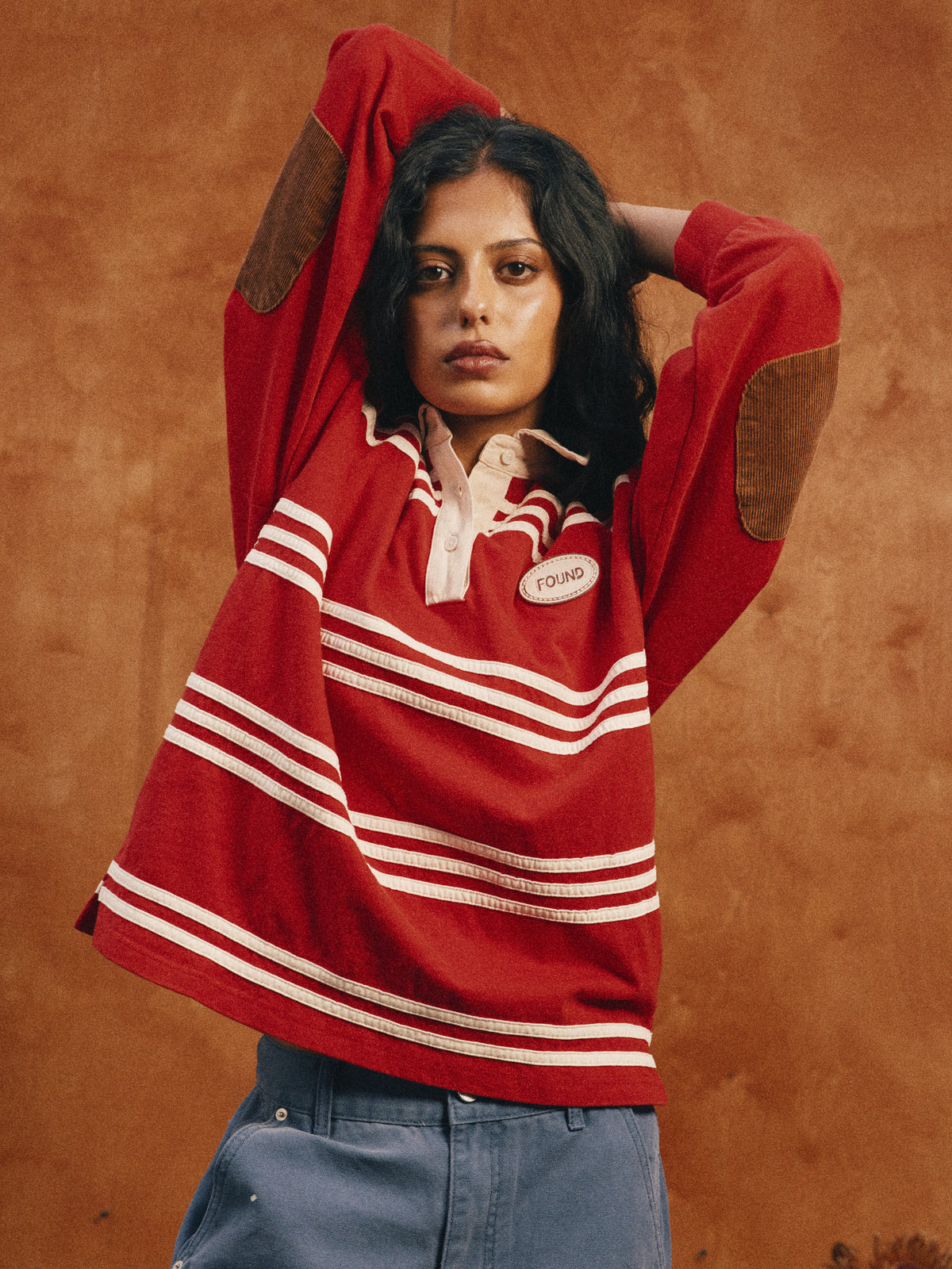 Someone wearing the FOUND Striped Corduroy Rugby Polo poses with arms raised behind their head against a brown background.