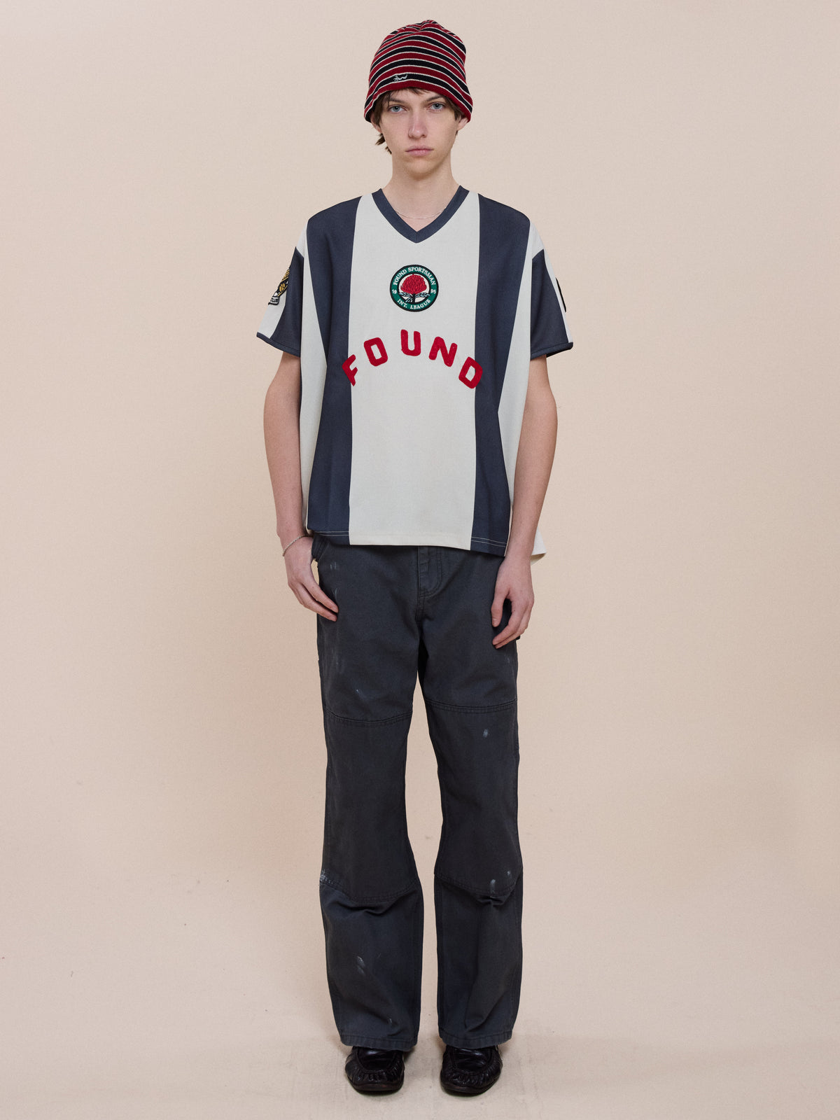Someone wearing the Striped Stateman Jersey by FOUND, a red striped beanie, dark pants, and black shoes stands against a plain beige background.