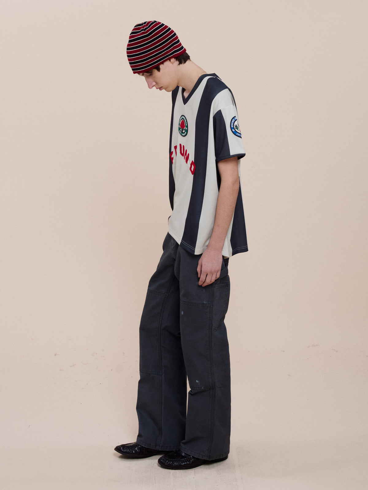 Person wearing a red-and-black striped beanie, FOUND Striped Stateman Jersey, dark wide-leg pants, and black shoes, standing and looking down against a plain background.