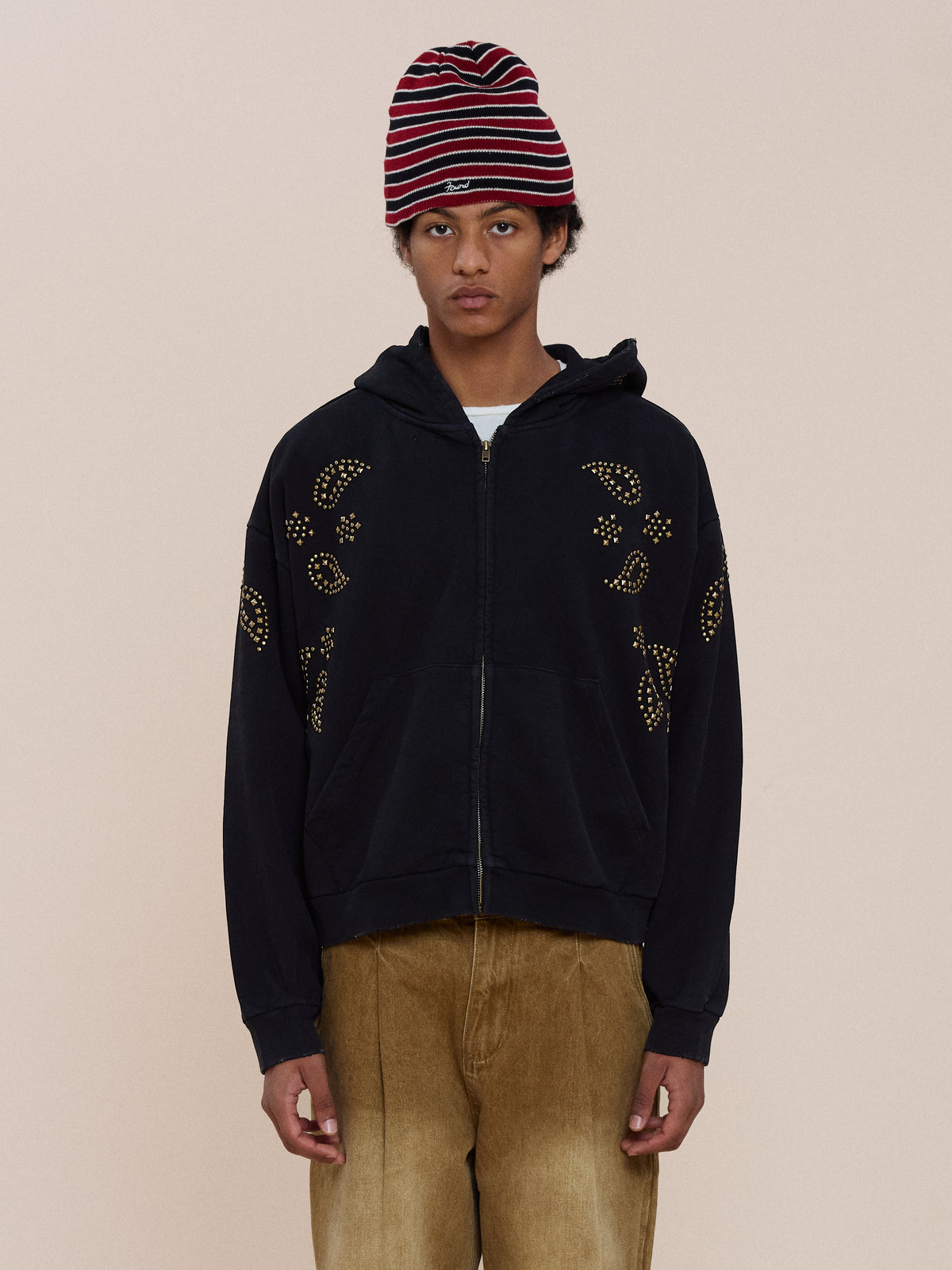 Person wearing the FOUND Studded Zip Up Hoodie, tan pants, and a red and white striped beanie, standing against a plain light background.