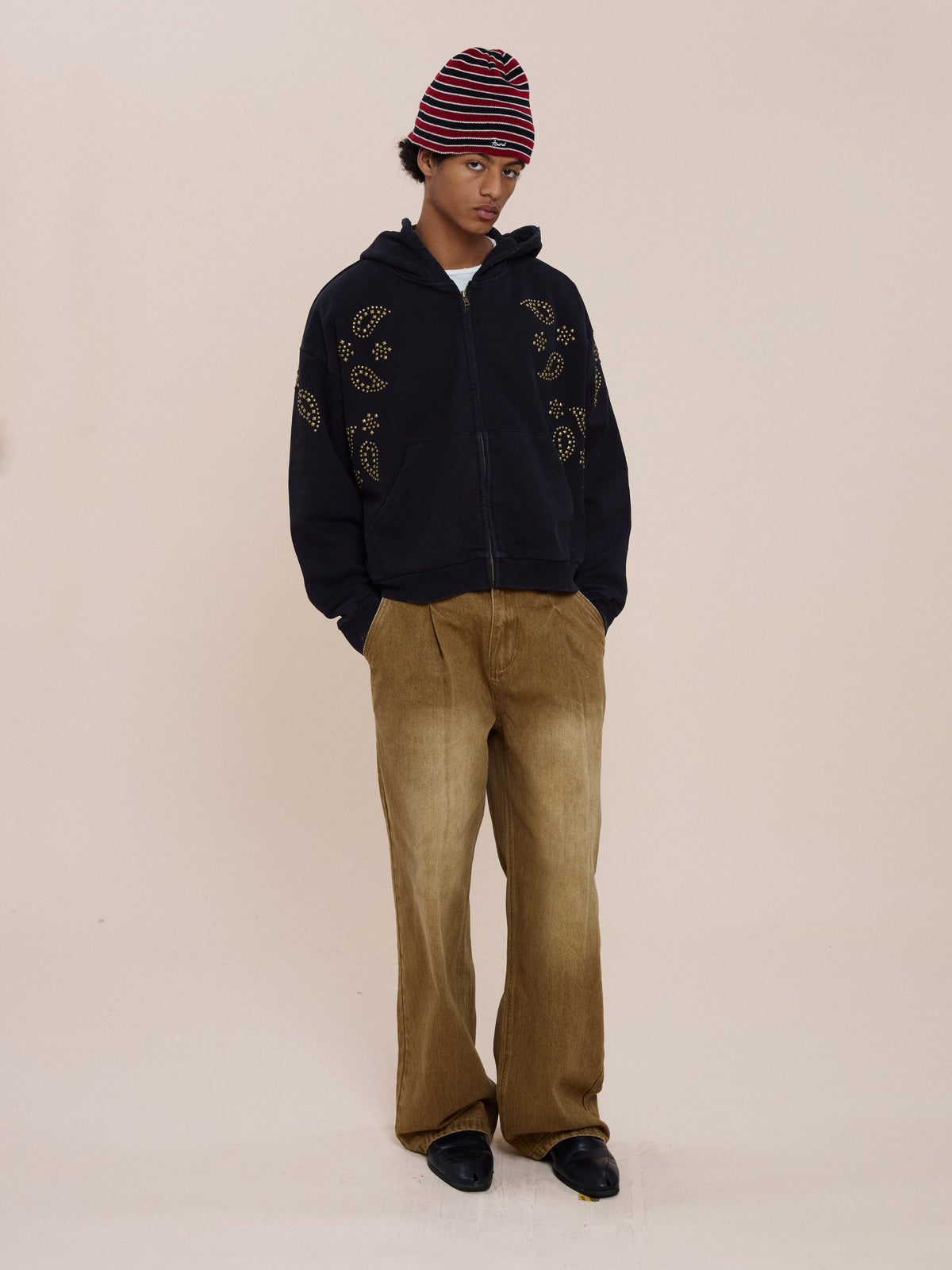 A person in a red and black striped beanie, FOUND Studded Zip Up Hoodie, wide brown pants, and black shoes stands against a plain beige background.
