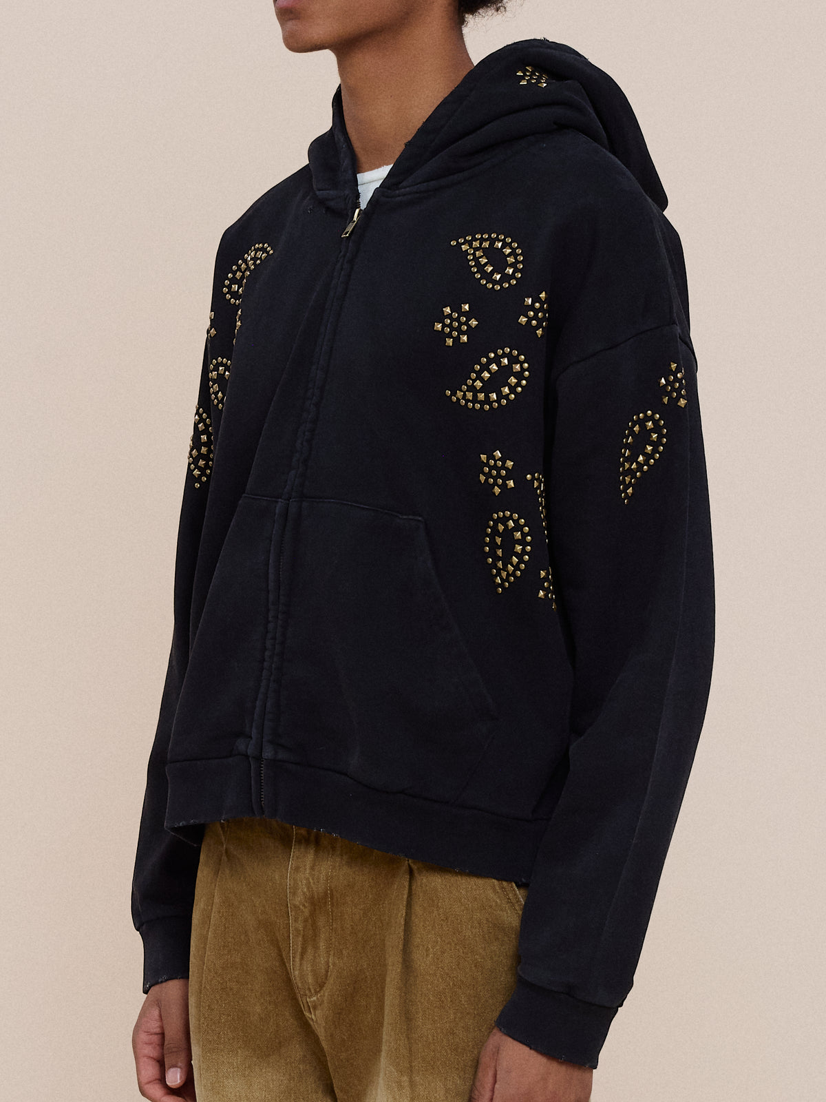 A person wears the FOUND Studded Zip Up Hoodie with gold studs and tan corduroy pants, standing against a plain light background.