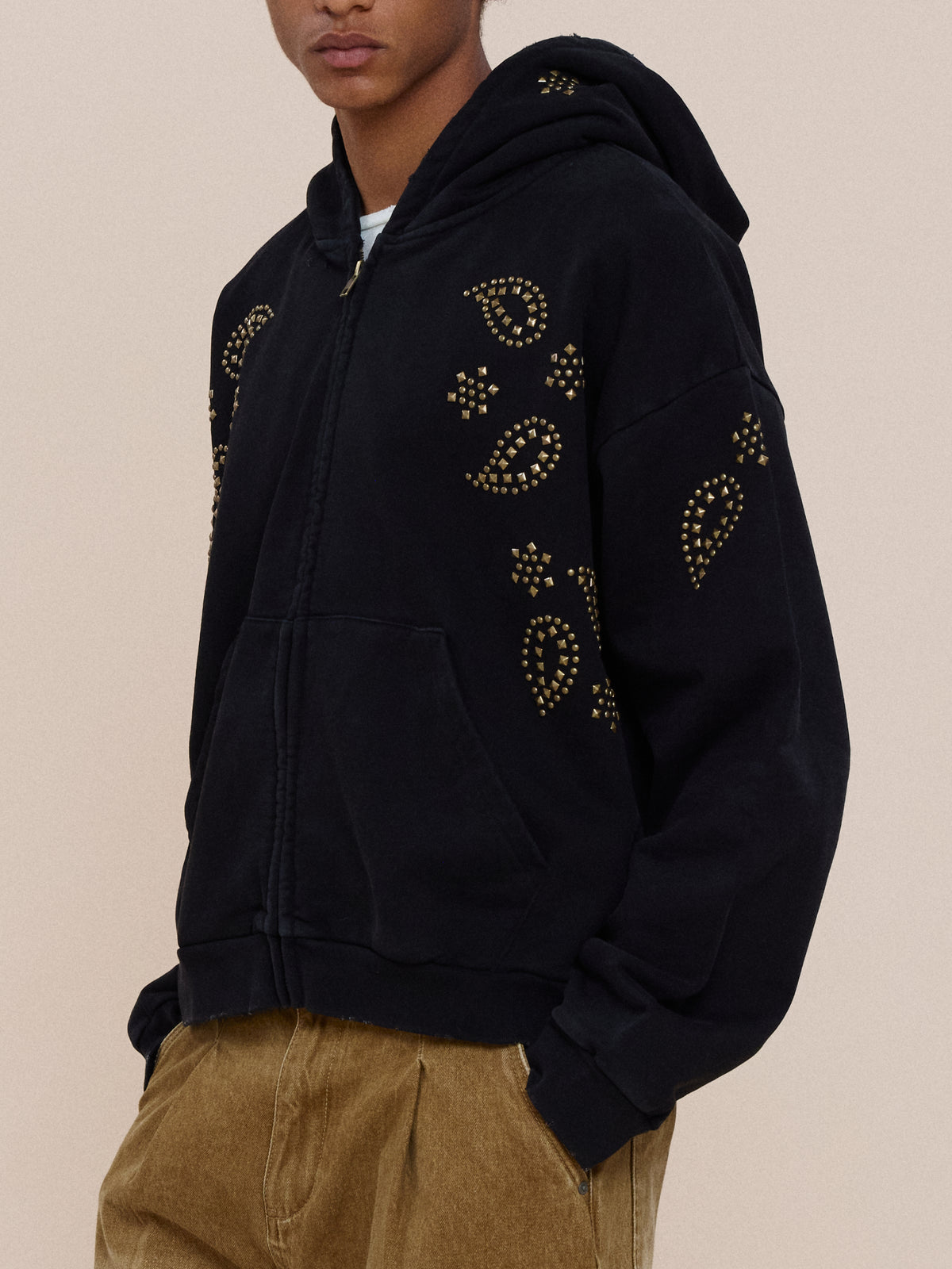 Model wears the Studded Zip Up Hoodie by FOUND with tan pants, standing hands-in-pockets against a light background.