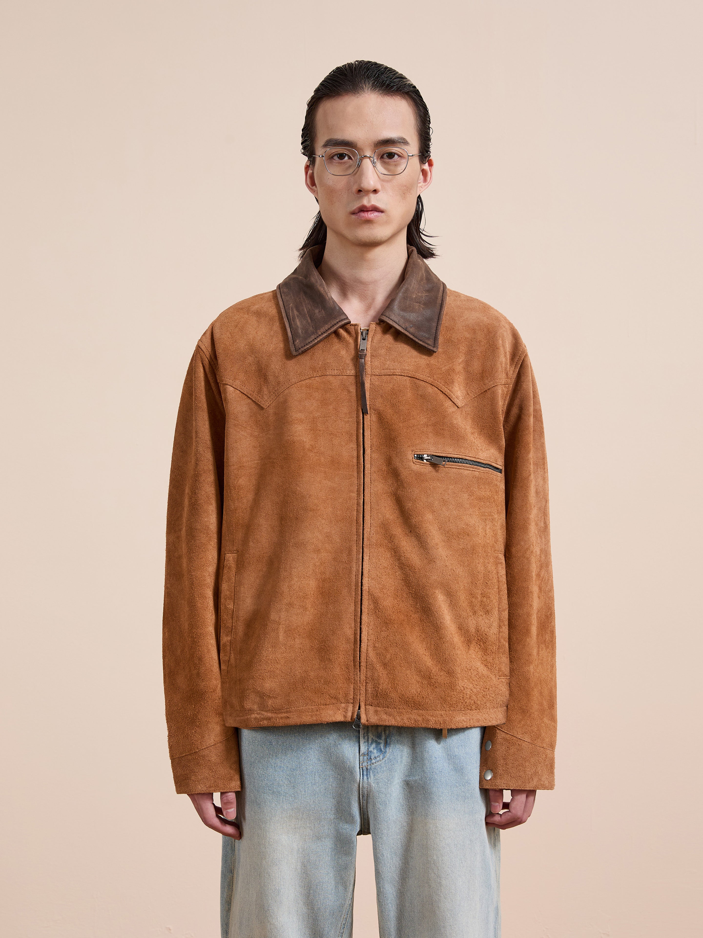 A person wearing glasses and the FOUND Suede Western Barn Jacket with a zipper pocket and western yoke, paired with light blue jeans, stands against a plain beige background.
