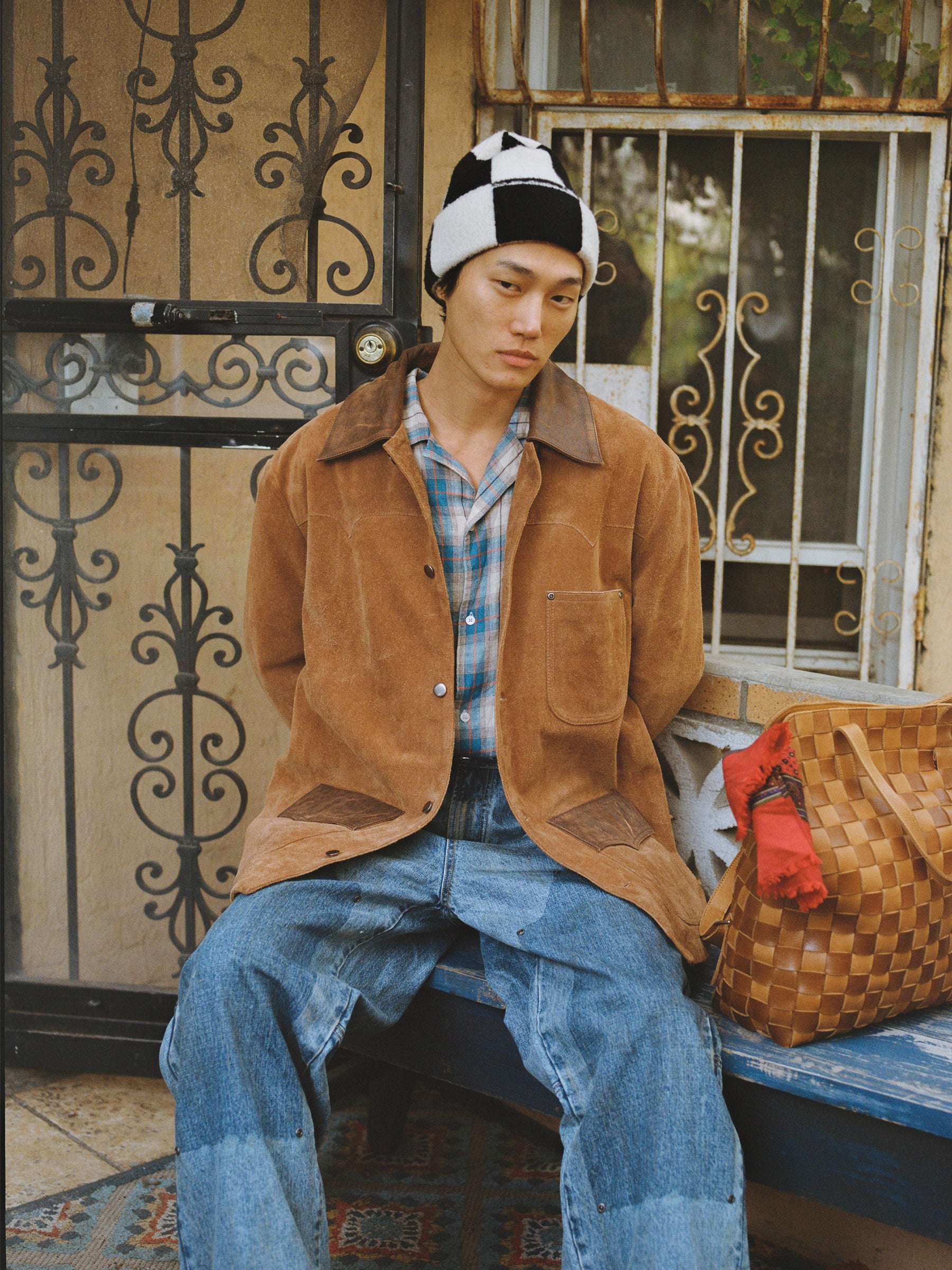 Wearing the FOUND Suede Contrast Chore Jacket, a plaid shirt, patchwork jeans, and a black-and-white beanie, someone sits on a bench beside a woven tote bag.