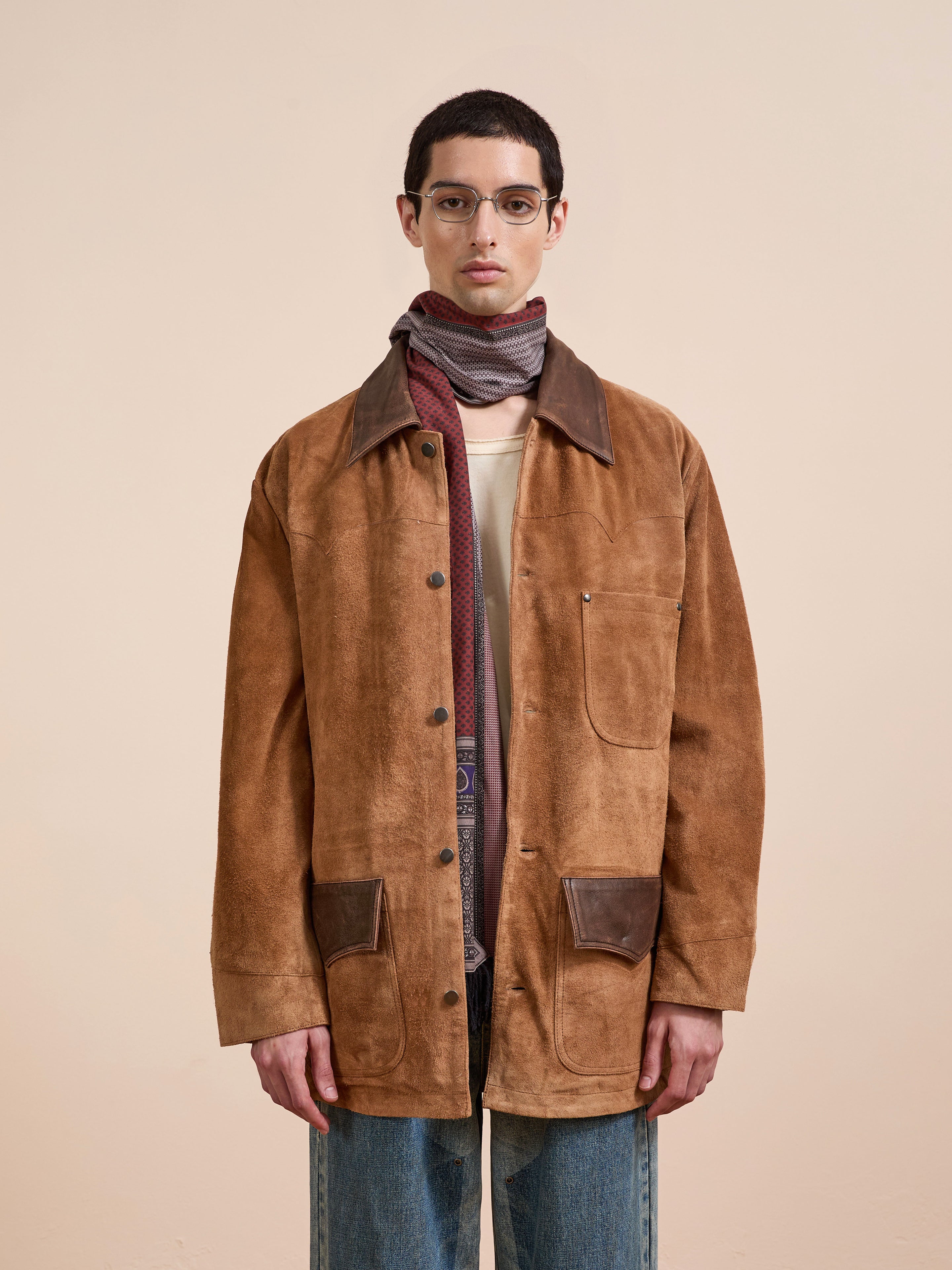 A person wears the FOUND Suede Contrast Chore Jacket with dark collar and pockets, styled with a scarf and blue jeans against a beige background. Its unisex relaxed fit ensures comfort in this chic look.