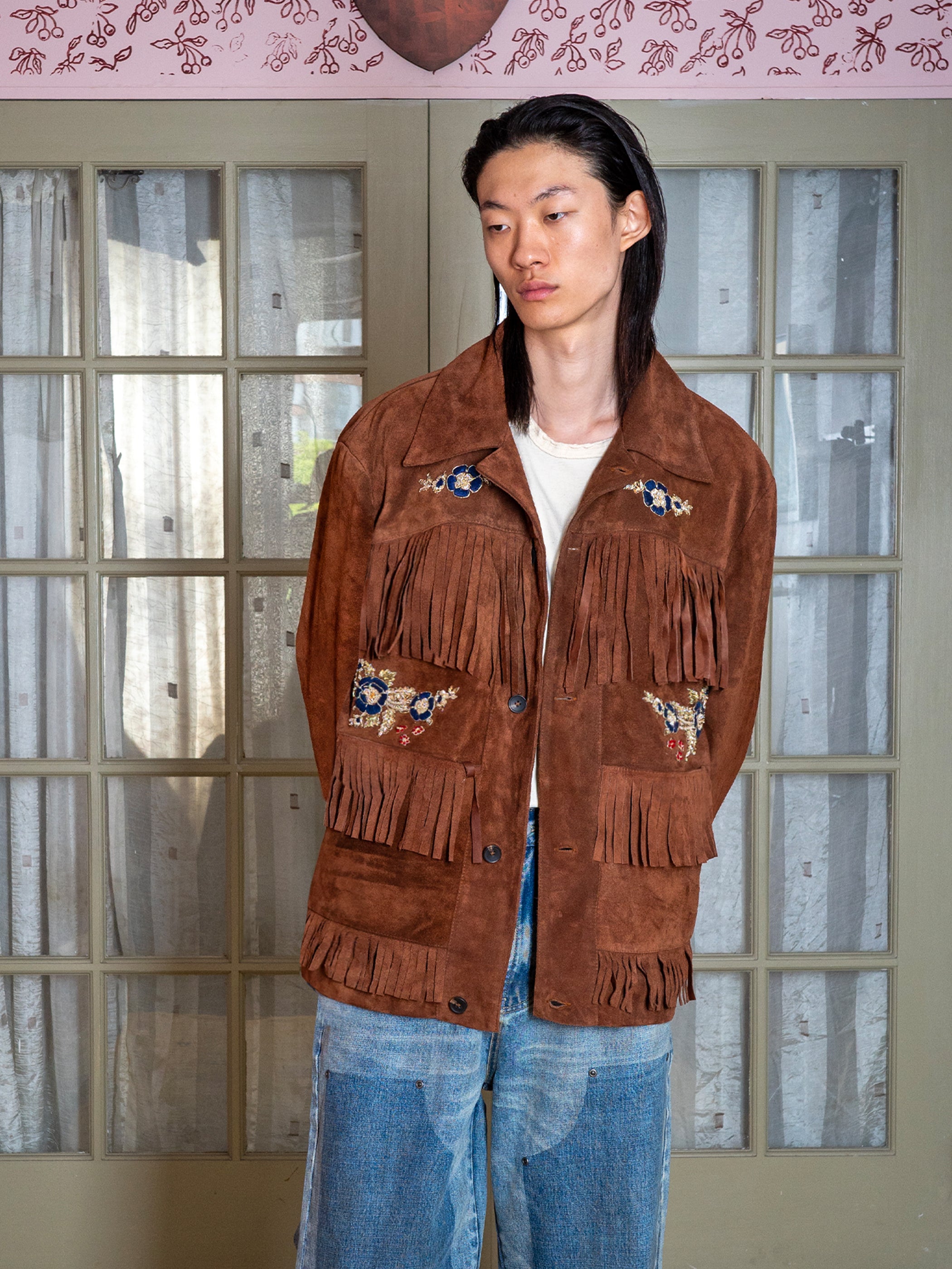 A person models the FOUND Hand Embroidered Suede Fringe Jacket over a white shirt and blue jeans, standing before glass-paneled doors draped with sheer curtains.