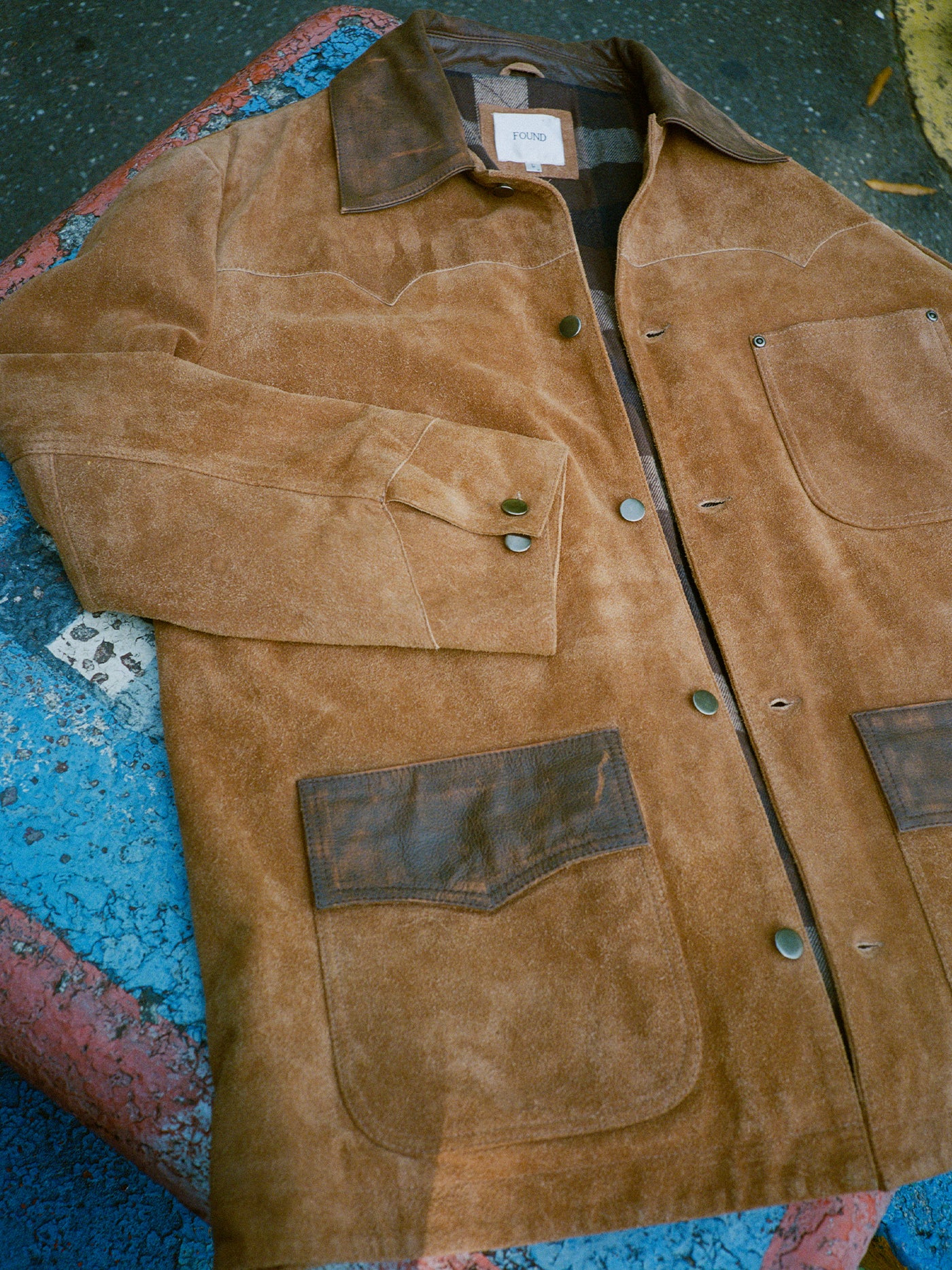 The FOUND Suede Contrast Chore Jacket features brown suede with dark brown leather collar and pocket accents, displayed flat on a weathered, multicolored surface for a timeless vintage appeal.
