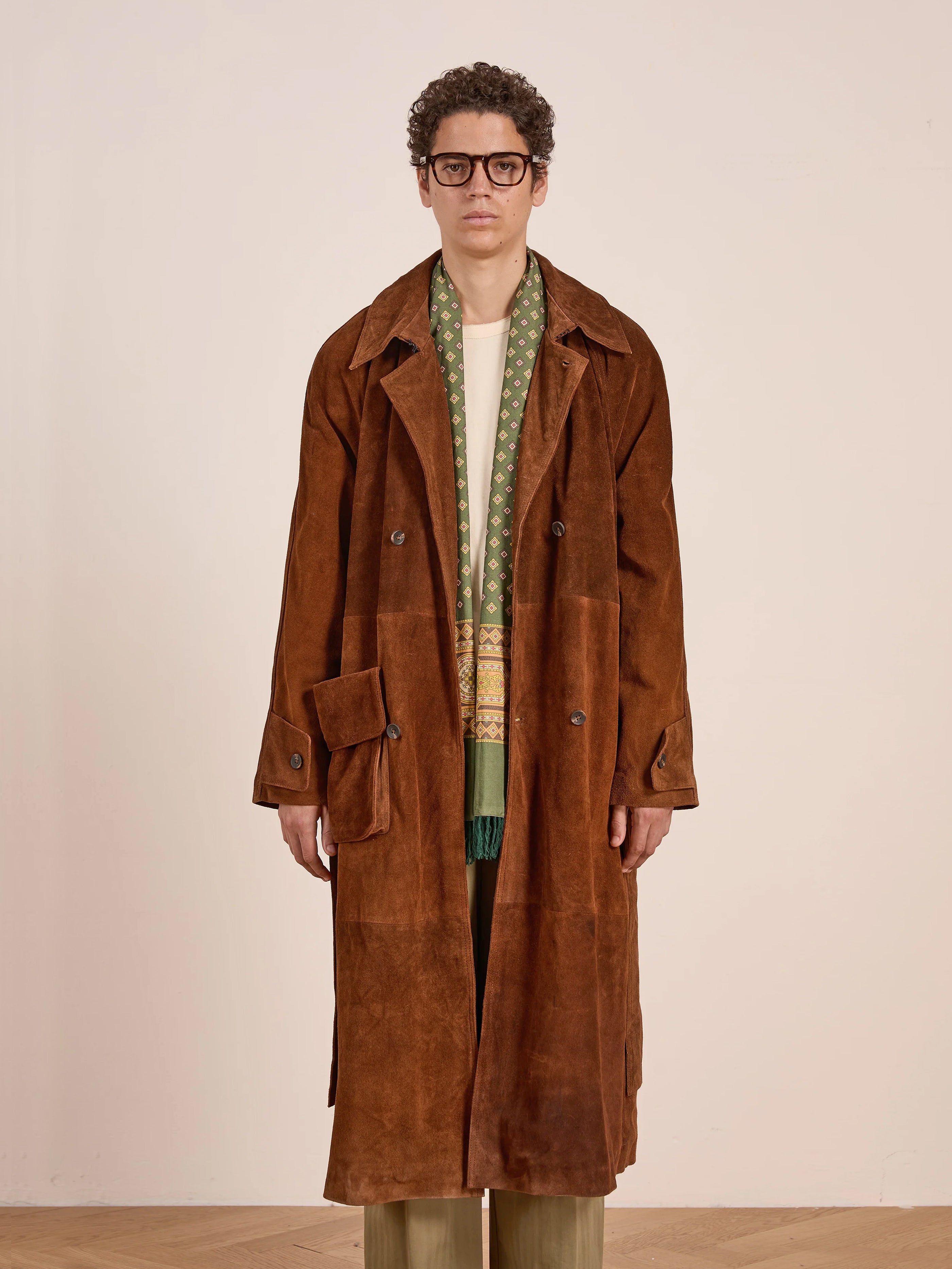 A person wearing glasses, a FOUND Suede Leather Trench Coat in luxurious brown cow suede, a patterned green scarf, and beige pants stands facing forward against a plain light background.