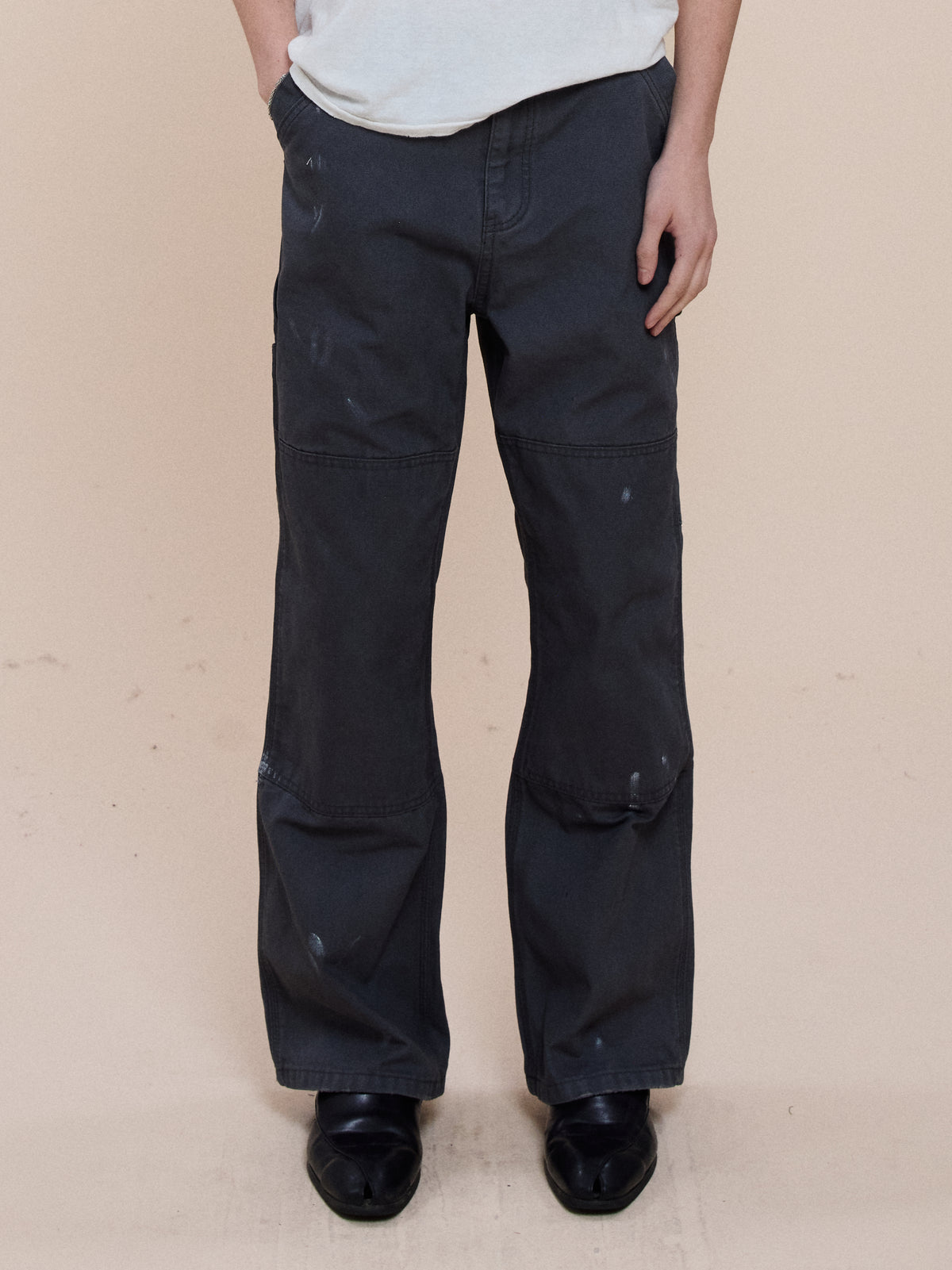 Model wears FOUND Sunbleached Utility Pants with paint marks, black shoes, and a white shirt against a beige background.