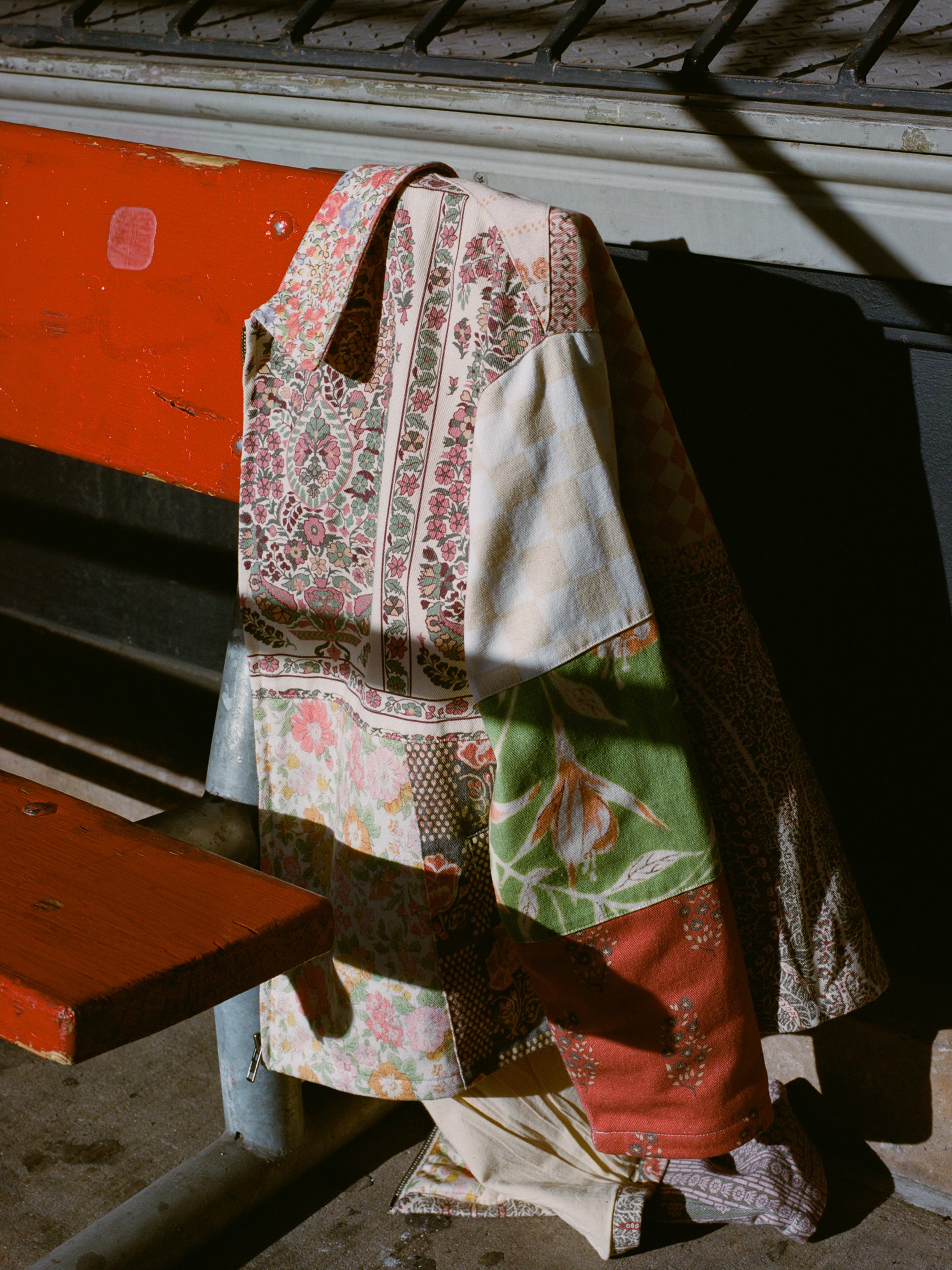 A patchwork quilt featuring floral and checkered patterns draped over a metal bench reflects the vintage style of FOUND's Tapestry Twill Jacket, while sunlight partially illuminates its wooden seat.