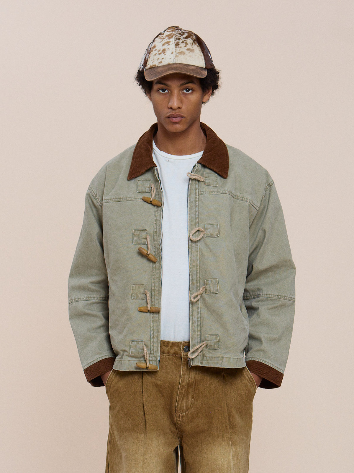 A person wears the FOUND Toggle Work Jacket—light denim with brown corduroy accents—paired with tan pants and a beige patterned cap, standing against a plain light background.