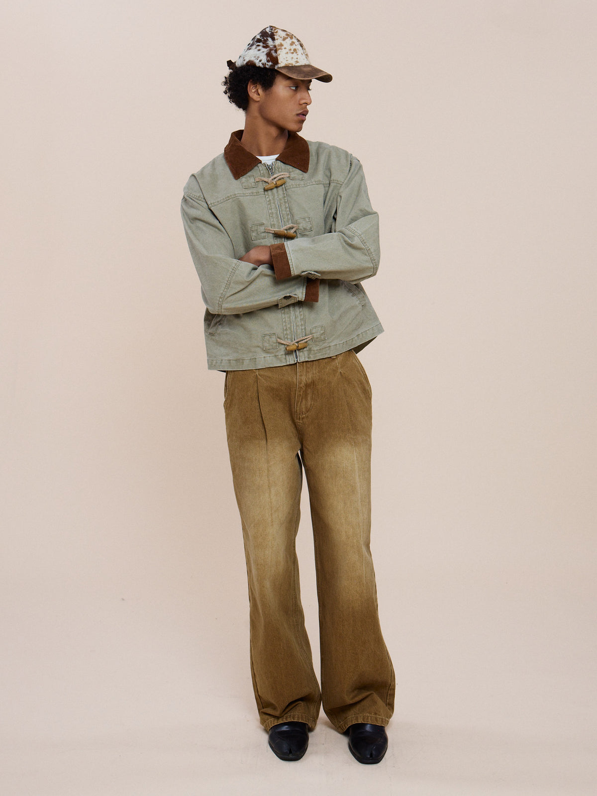 A person in the light green FOUND Toggle Work Jacket with brown accents, wide tan pants, and a patterned cap showcases vintage utility outerwear style.
