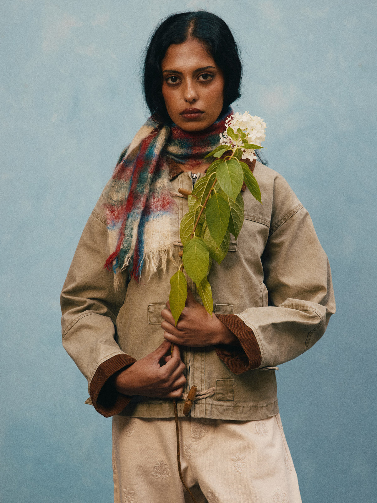 A person wearing the Toggle Work Jacket by FOUND stands against a blue background, holding a leafy stem with a white flower.