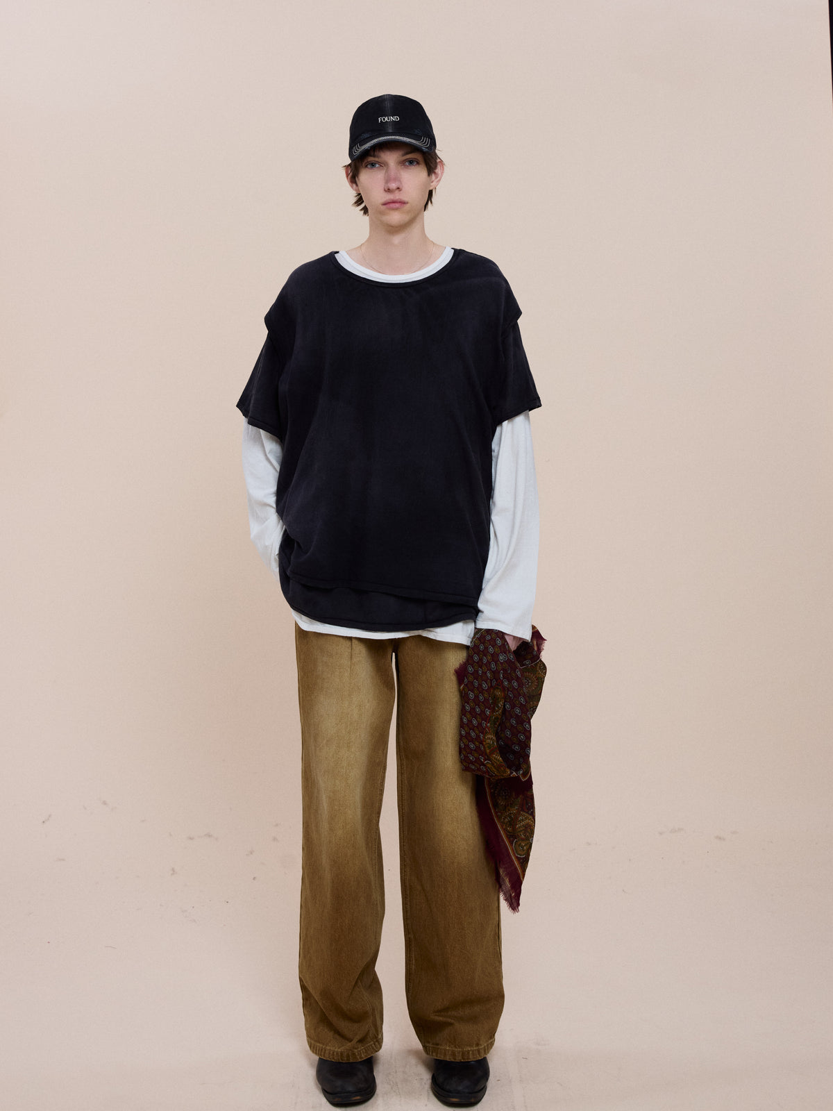 Person stands against a beige background wearing FOUND’s Triple Layer Tee, black cap, wide brown pants, black shoes, and holding patterned fabric.