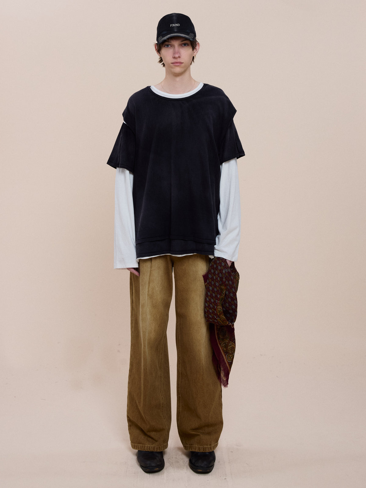 Person in black cap, FOUND Triple Layer Tee over white long-sleeve, wide brown pants, black shoes, and patterned scarf stands against a plain beige background.