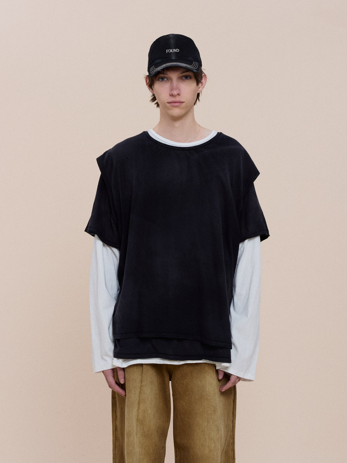 Person stands against a beige background wearing the FOUND Triple Layer Tee in black over a white long-sleeve, paired with loose tan pants and a black cap.