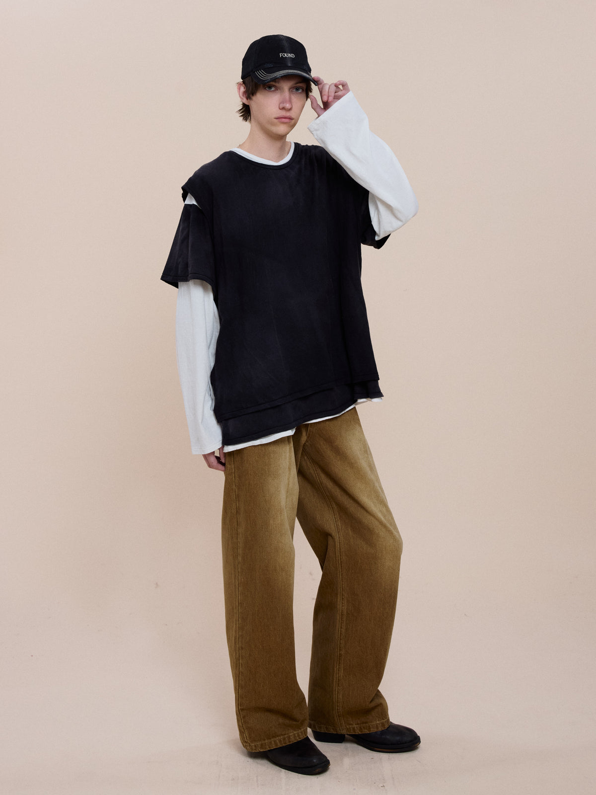 A person in a black cap, FOUND Triple Layer Tee (unisex relaxed fit), wide brown pants, and black shoes stands against a plain beige background.
