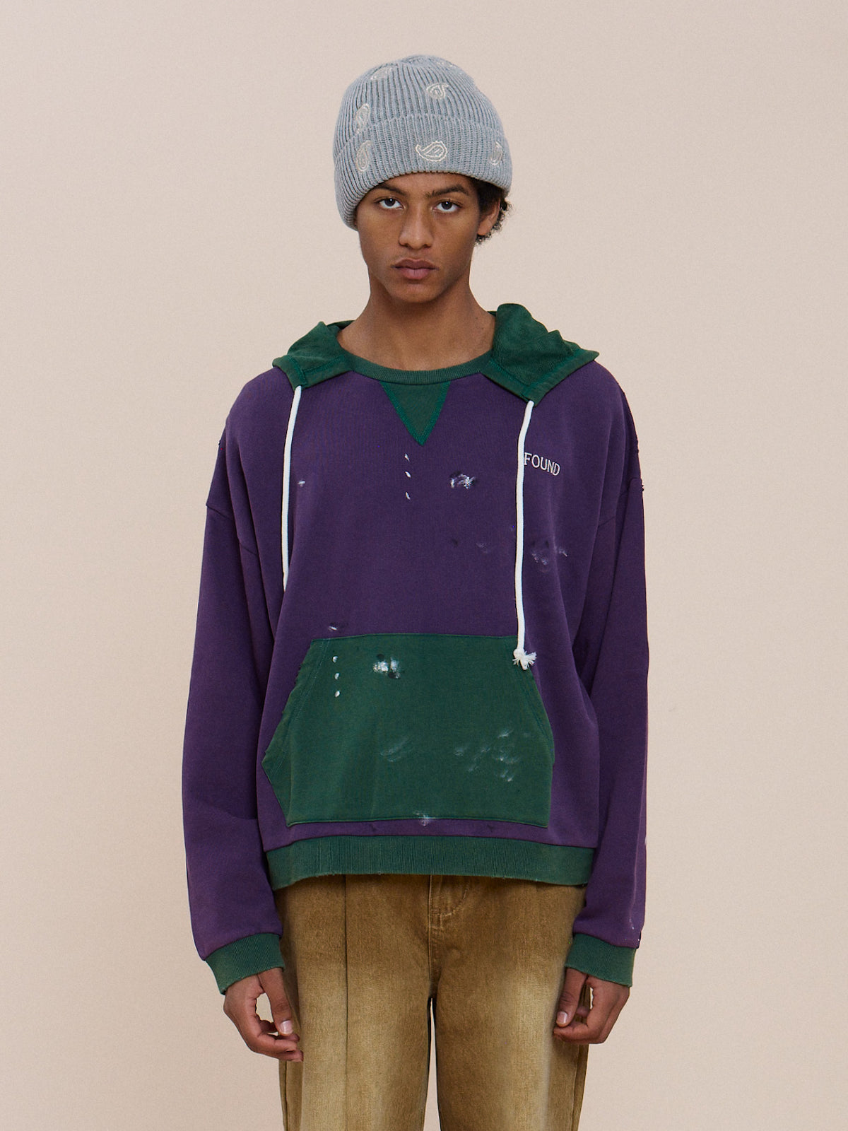 A person in a gray knit beanie, tan pants, and the FOUND Two Tone Painter Hoodie stands against a plain beige background.