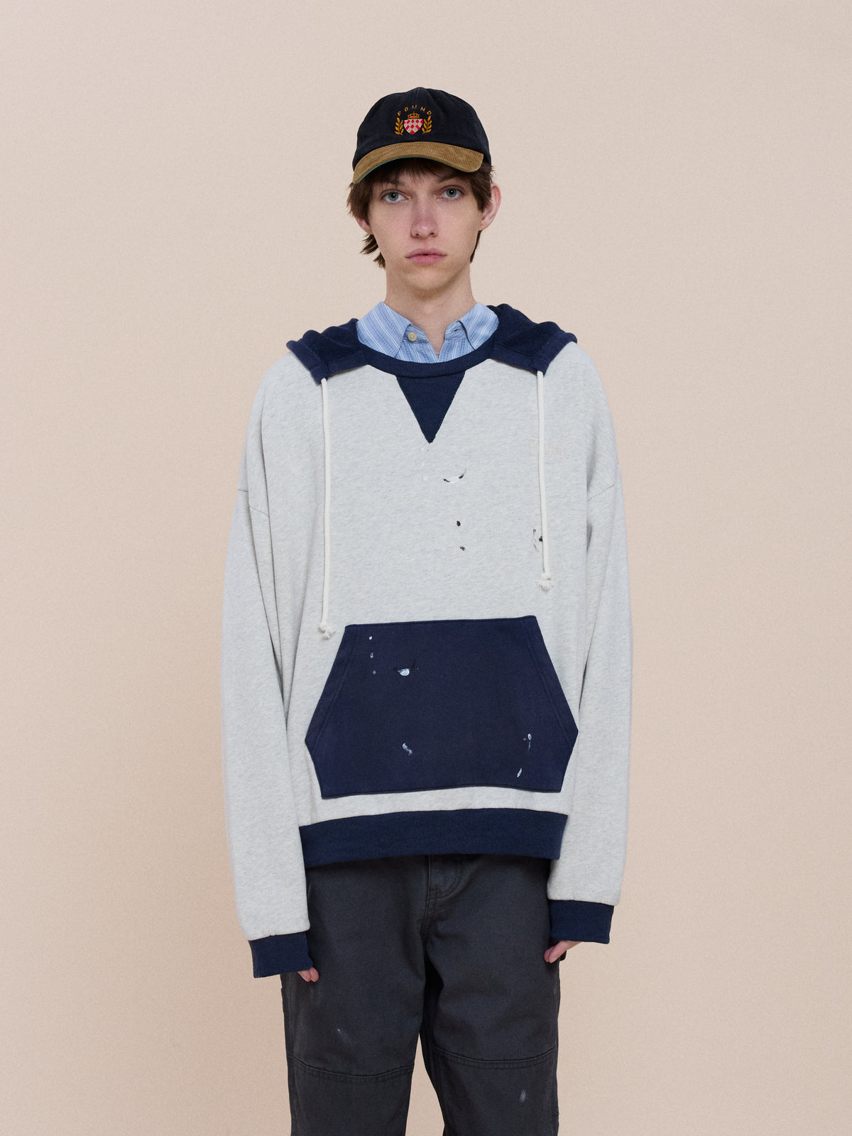 Someone stands in front of a plain beige background, wearing the FOUND Two-Tone Painter Hoodie over a collared shirt, baseball cap, and dark pants.