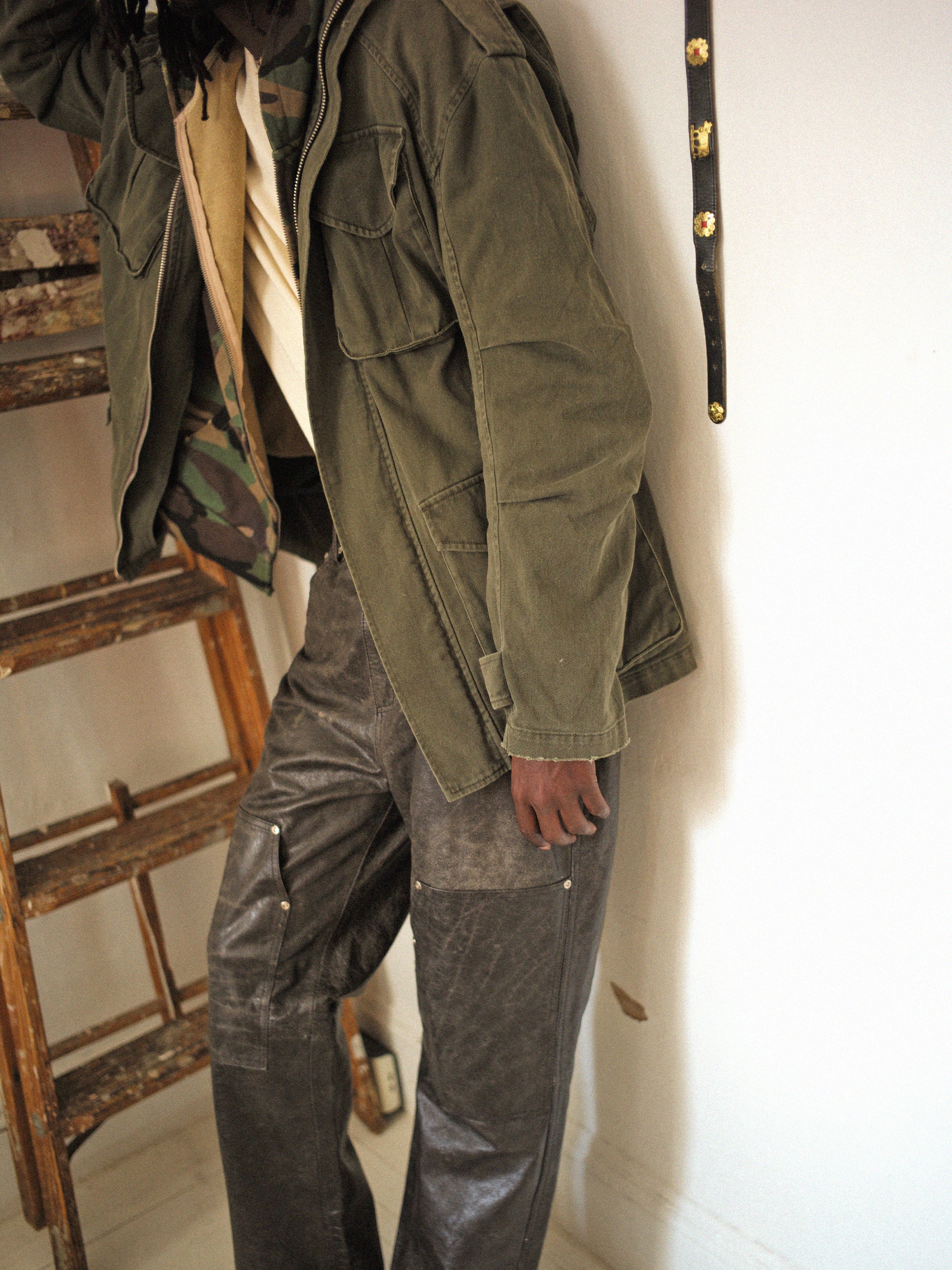 A person in an olive green M65 Utility Twill Pocket Jacket by FOUND and dark pants leans against a wooden ladder next to a white wall, showcasing a vintage look with military-inspired outerwear.