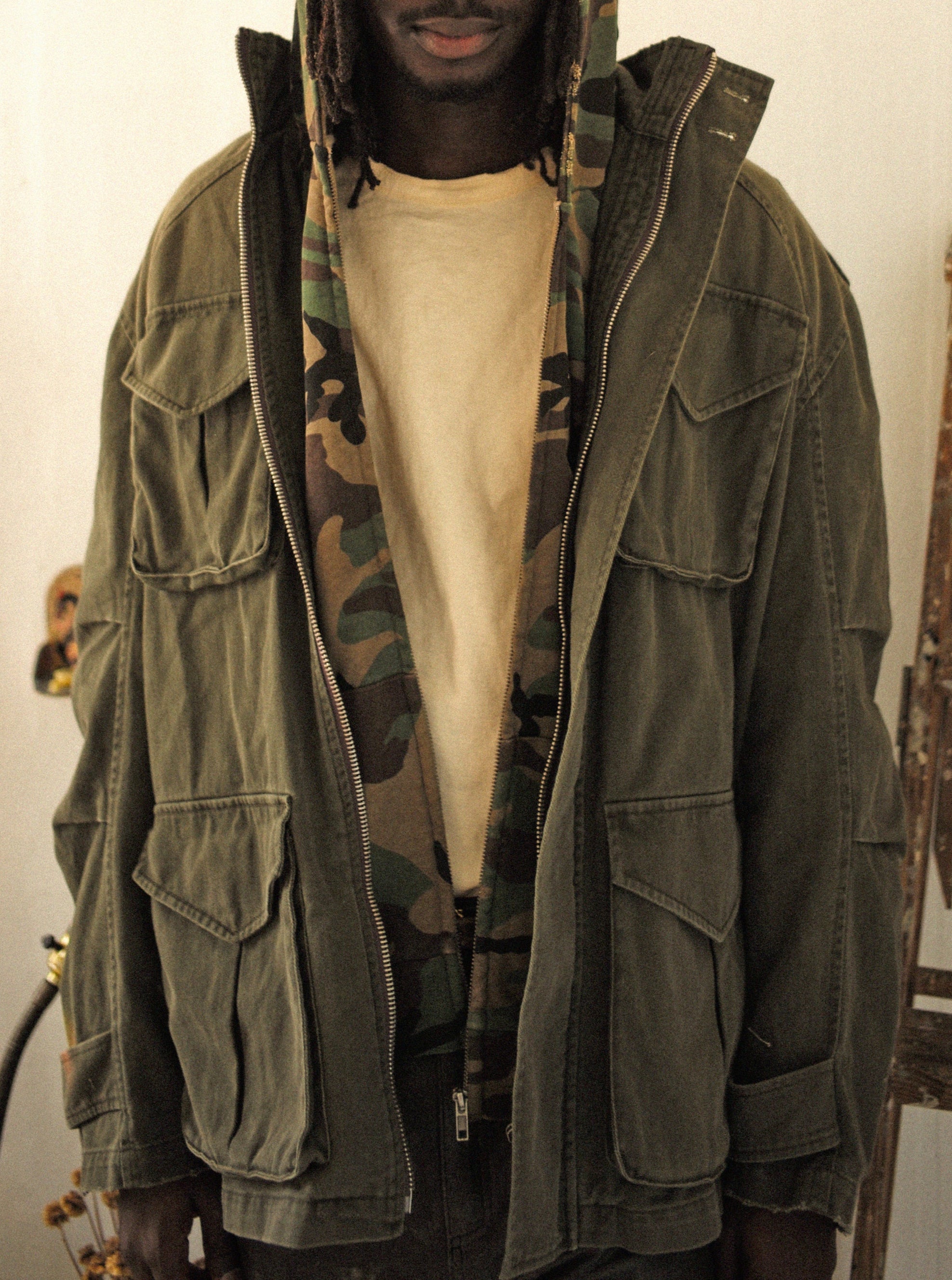 A person is indoors wearing a FOUND M65 Utility Twill Pocket Jacket in green over a camouflage hoodie and yellow shirt.