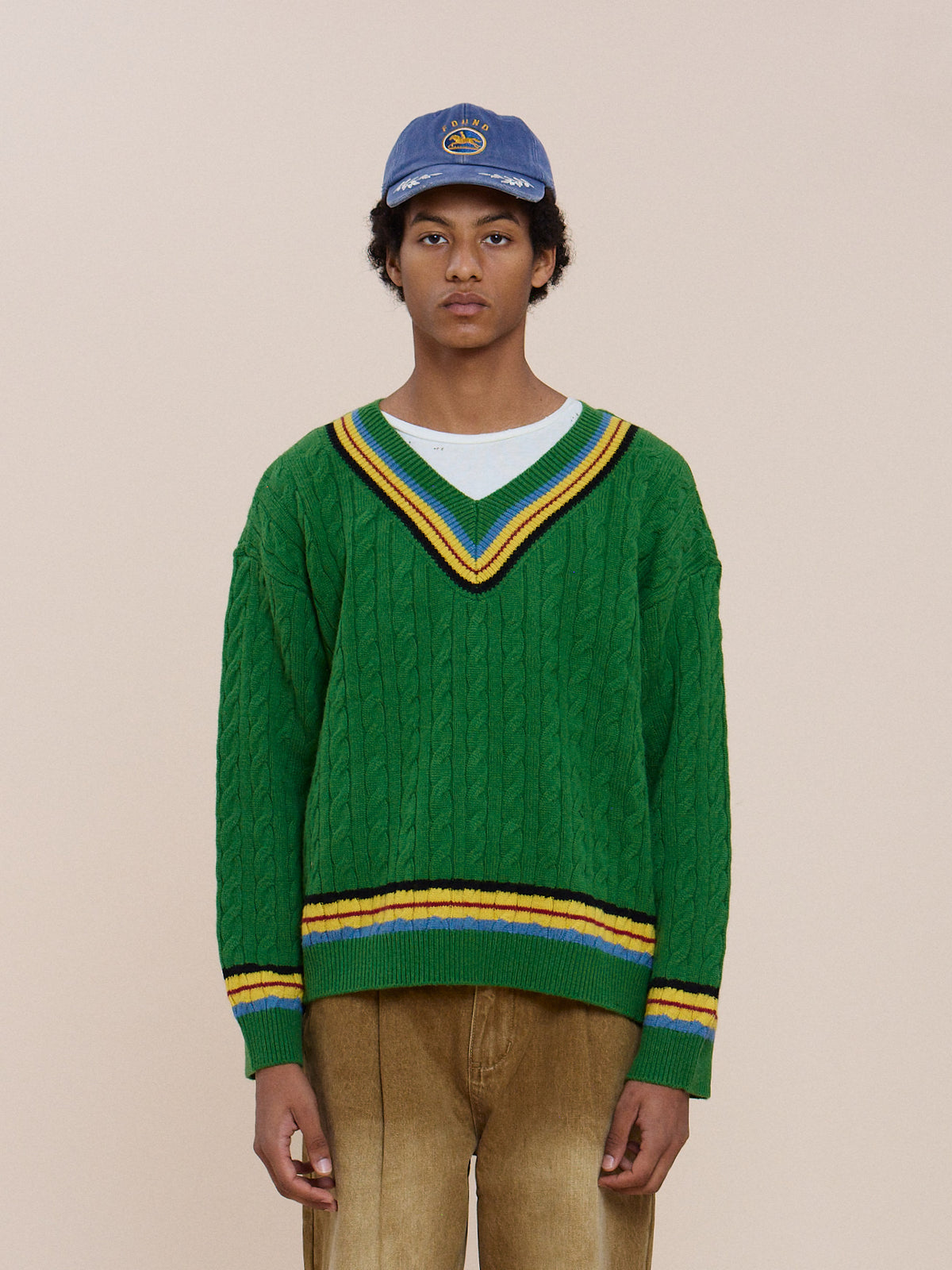 A person wears the FOUND V-Neck Cricket Sweater (green with yellow and black stripes), a white shirt, tan pants, and a blue cap against a beige background.