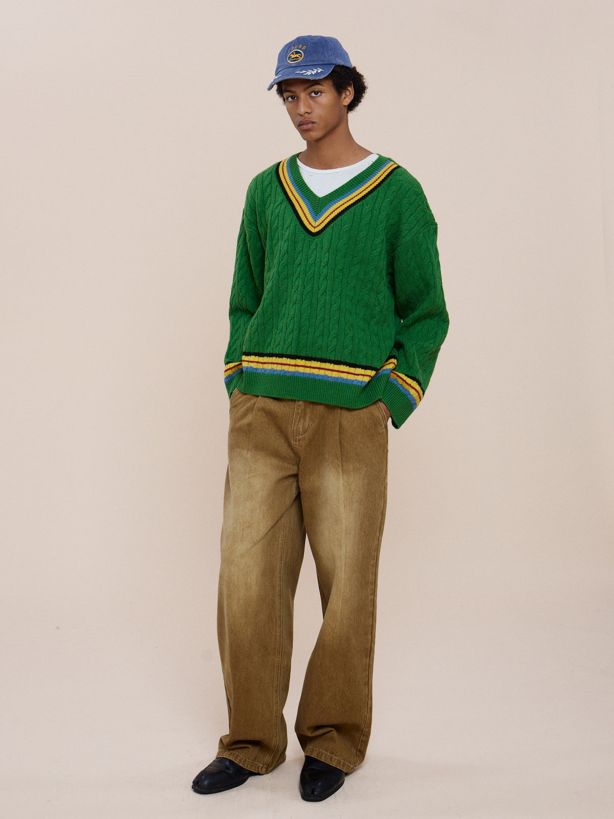 A person in a blue baseball cap, FOUND V-Neck Cricket Sweater with yellow stripes, brown wide-leg pants, and black shoes stands against a beige background.