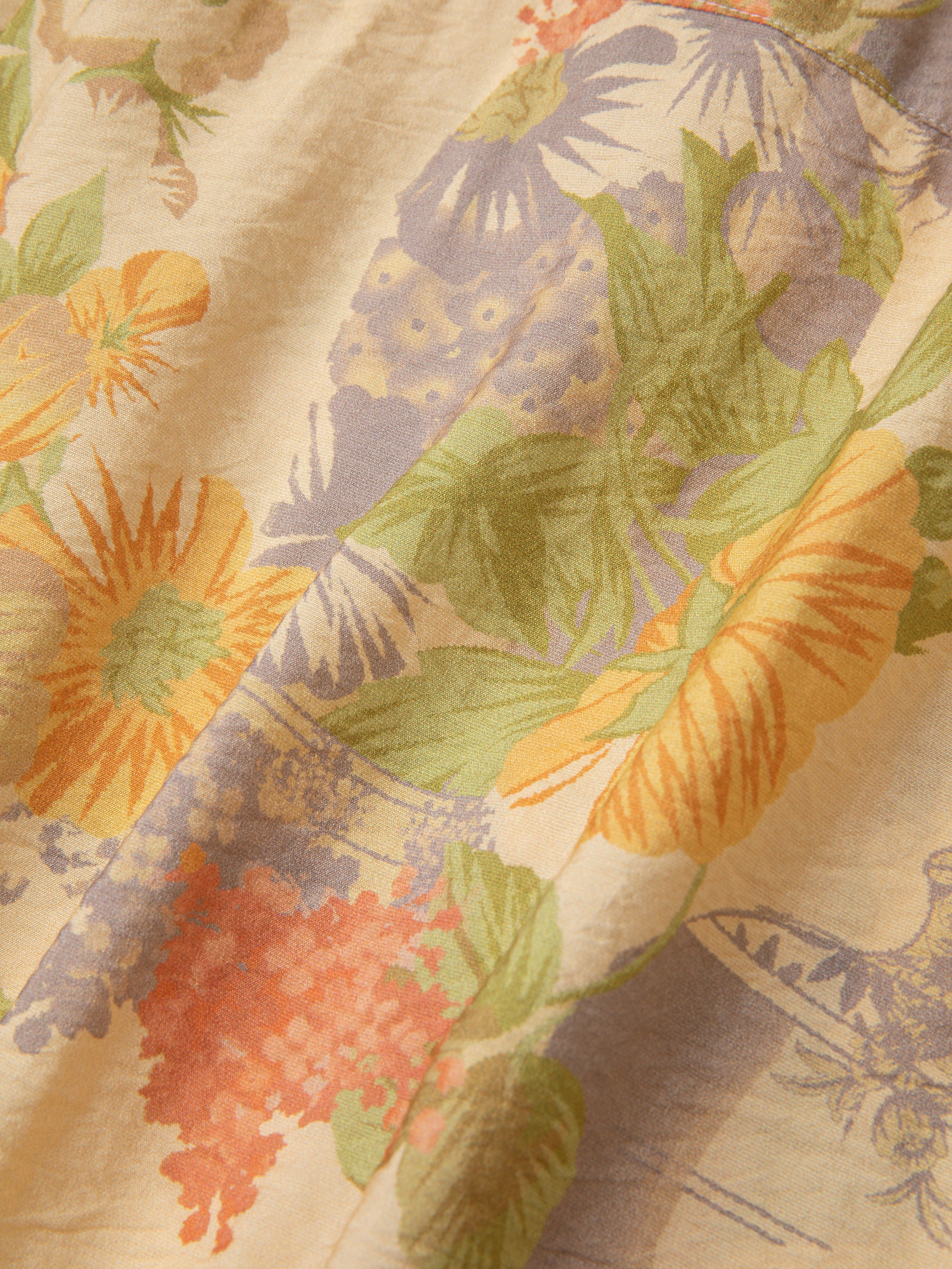 Close-up view of a fabric with a floral pattern blending sunflowers, leaves, and various other flowers in shades of yellow, orange, and green on a light background, reminiscent of traditional Phulkari motifs from Punjab Pakistan. The fabric is part of the Meraj Vase Pot Long Sleeve Camp Shirt by Found.