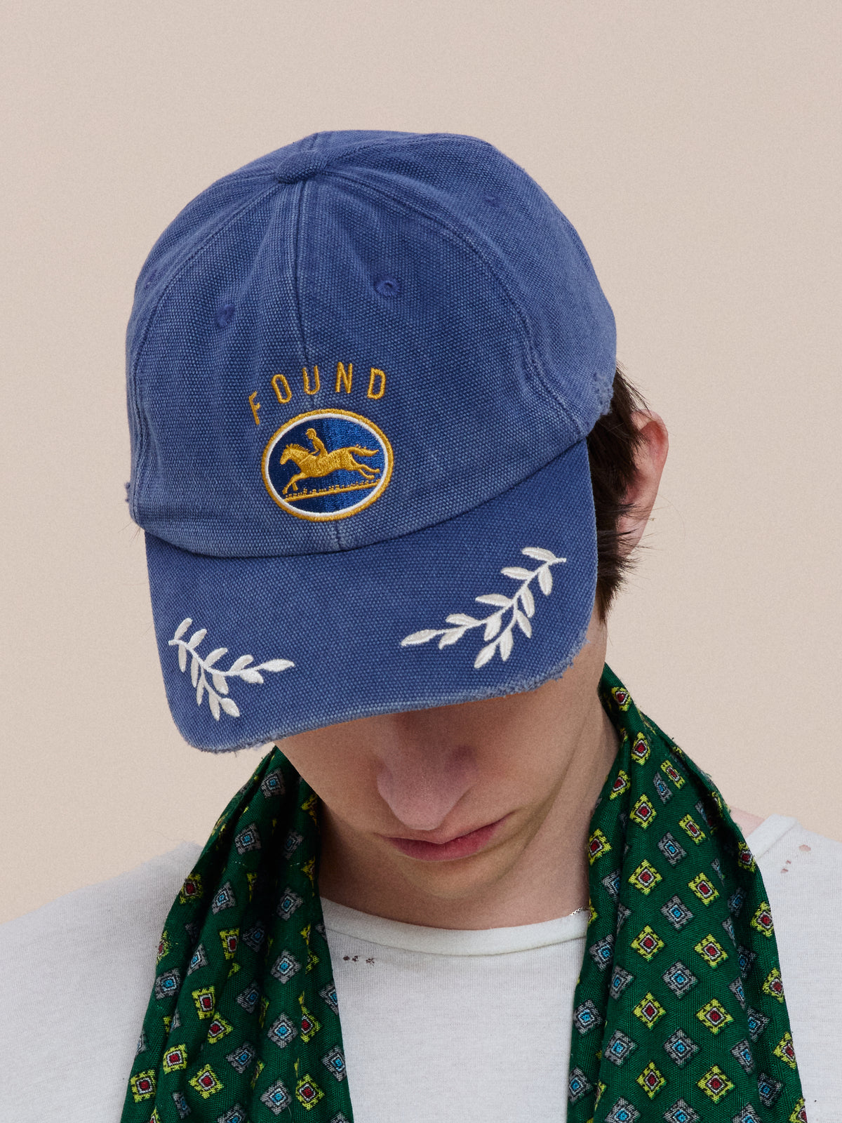 A person looks down, wearing the Vintage Club Cap by Found—blue with a yellow “FOUND” logo, white leaf details, an adjustable leather strap, and a green patterned scarf.