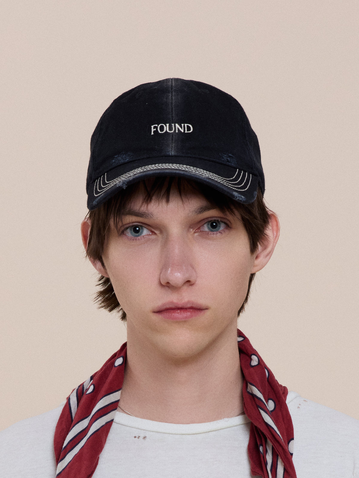 A person with straight brown hair wears the Washed Logo Cap by Found, a red patterned scarf, and a white shirt, facing forward against a plain background.