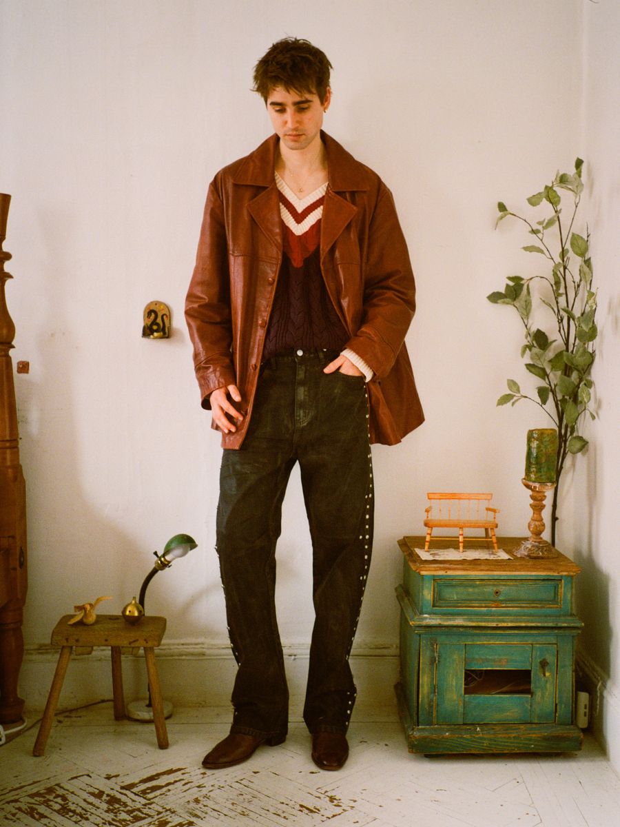 A person wearing FOUND's Waxed Studded Jeans and a brown waxed cotton jacket stands in a room with a wooden chair, decor on a small table, and a plant.