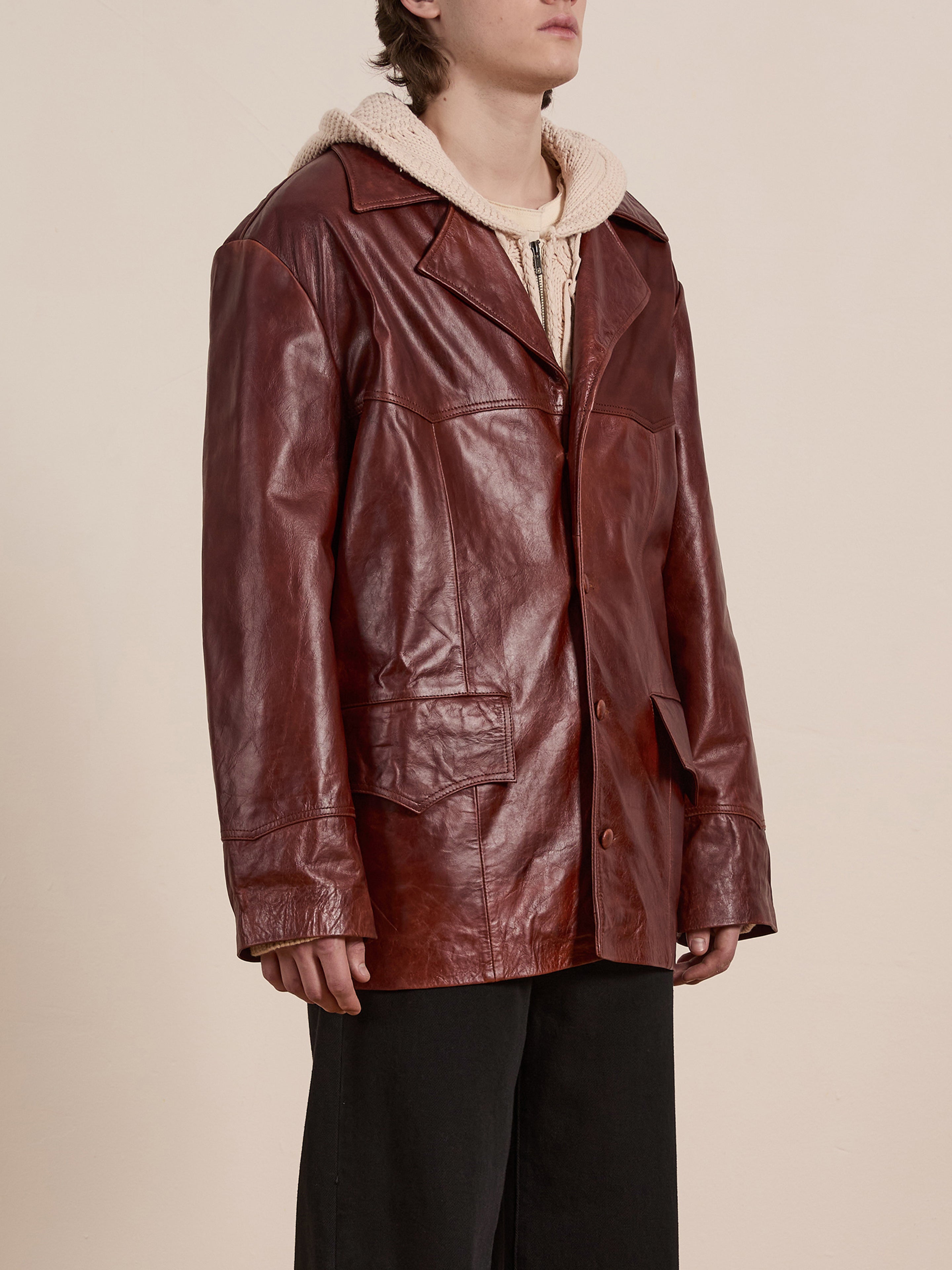Sporting a chic unisex look, they effortlessly pair wide black pants with a FOUND Claret Western Oversized Leather Blazer over a beige hoodie. The plain background accentuates their fashionable flair.
