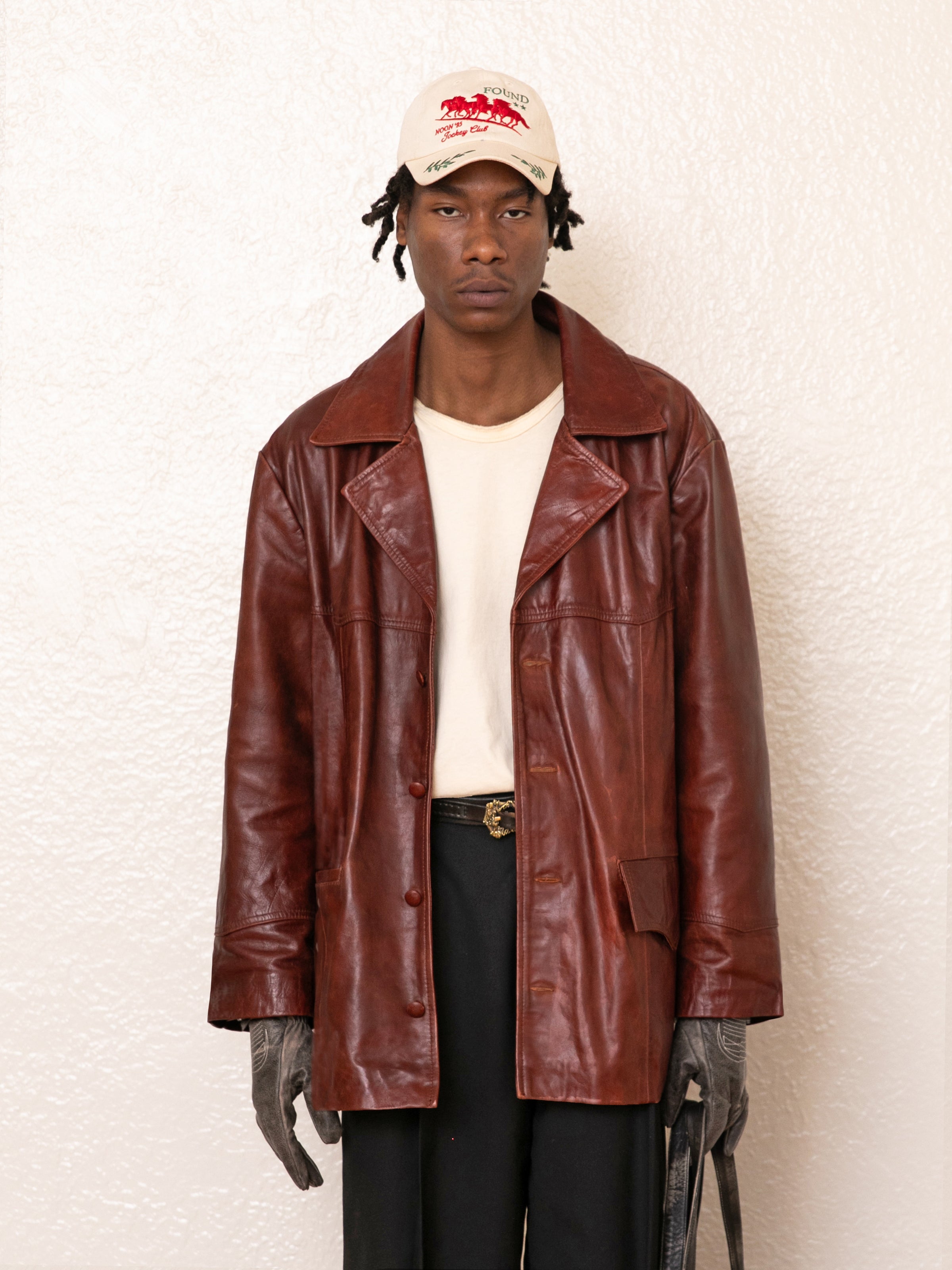 Clad in a FOUND Claret Western Oversized Leather Blazer over a cream shirt, with black pants, gloves, and a beige cap, the person stands against a textured white wall.