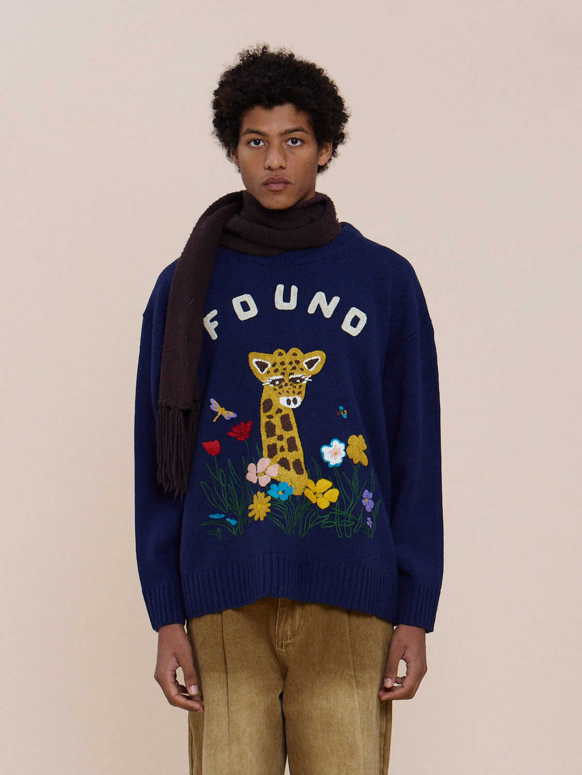 Person wearing the FOUND Wildlife Knit Sweater with giraffe graphic, tan pants, and dark scarf, standing against a beige background.