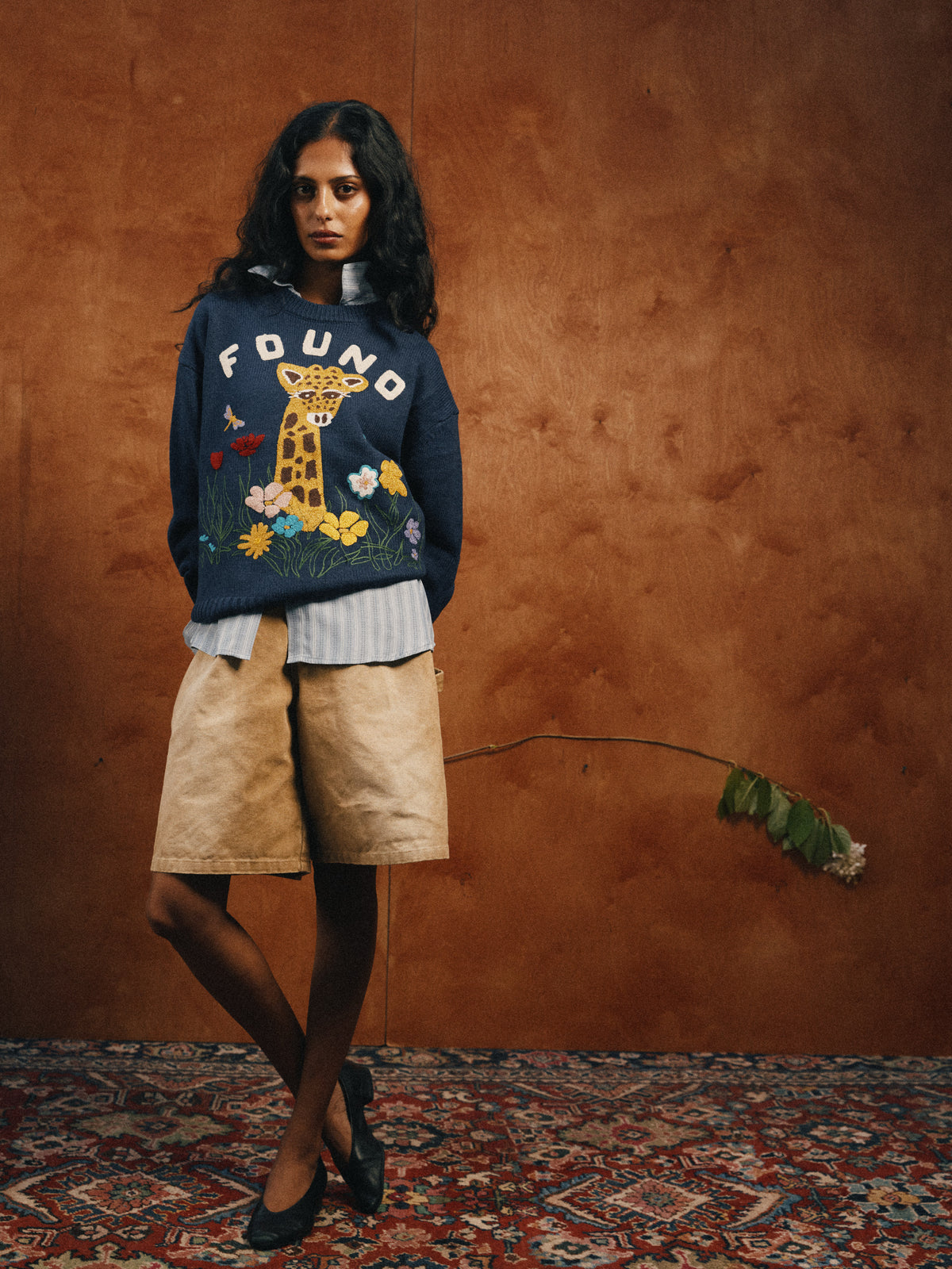 Woman stands indoors by a wood wall, wearing the FOUND Wildlife Knit Sweater with giraffe graphic, a striped shirt, tan shorts, and black shoes.